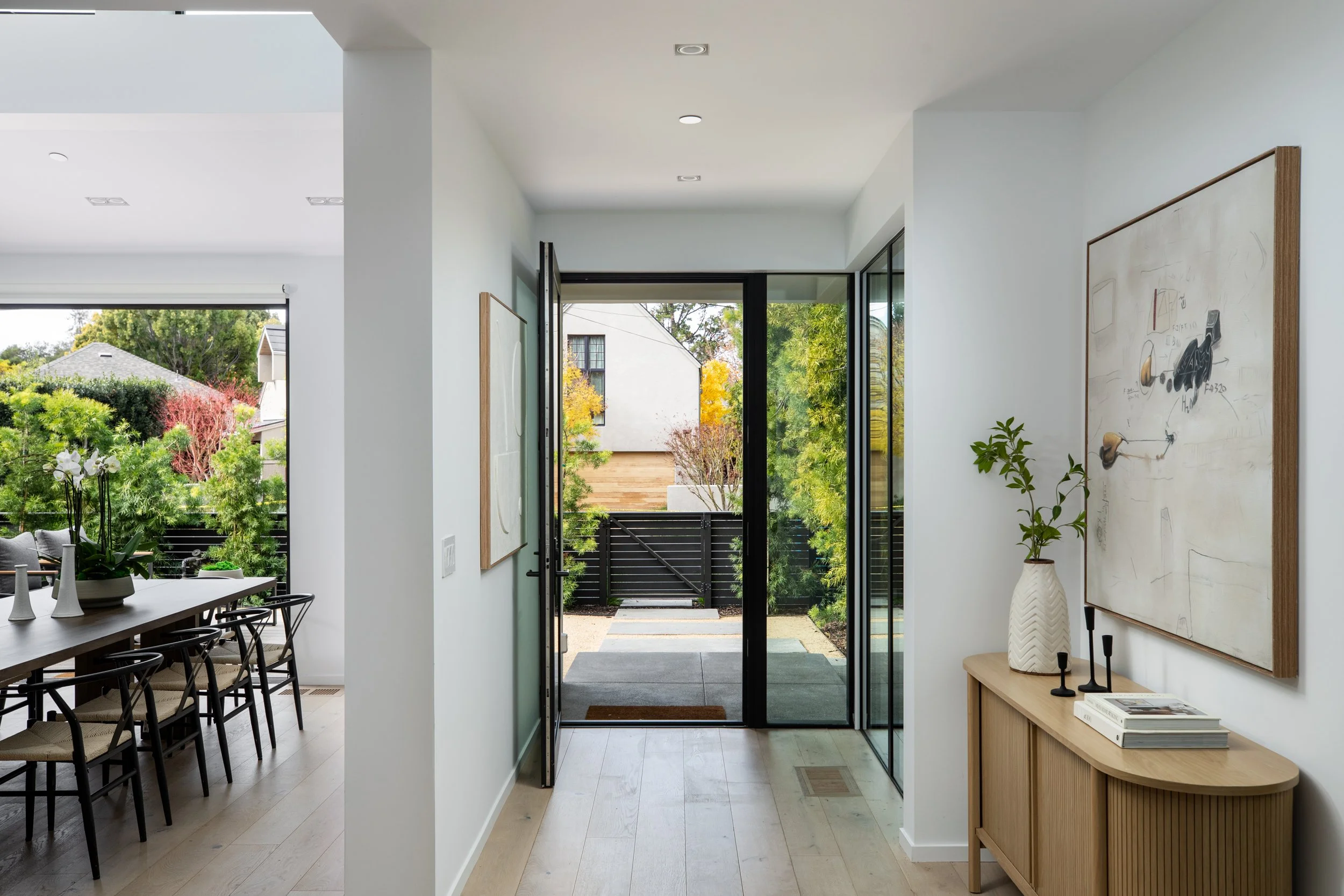Entryway of a modern home with an open glass door leading to a backyard with trees and a black fence, and a dining area with black chairs and a dark table on the left.