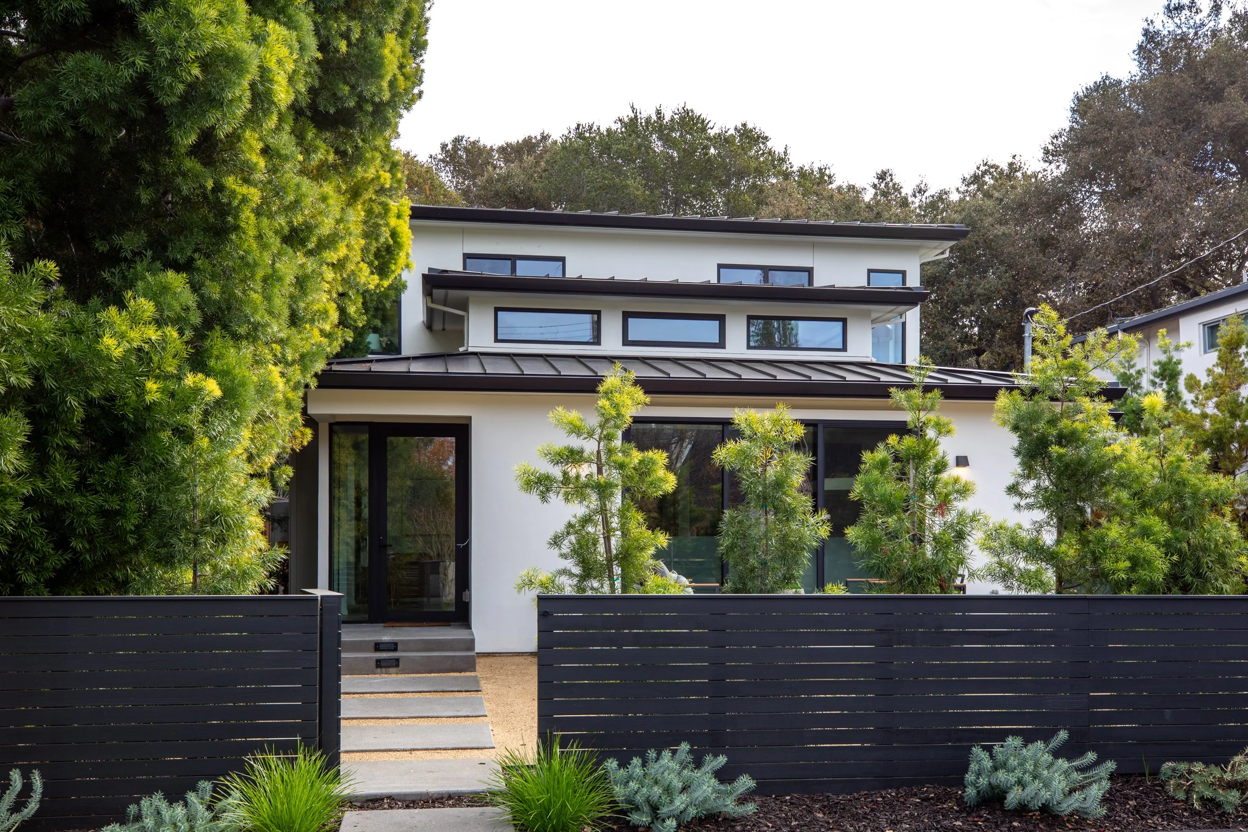 Modern white multi-story house with black window frames, surrounded by green trees and shrubs, with a black fence in front.