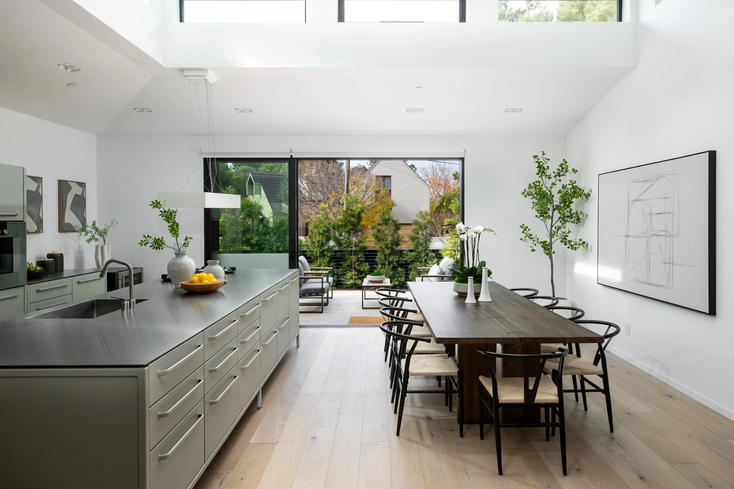 Modern open-plan kitchen and dining area with a view of a patio and trees outside, featuring a large central island with a faucet, a wooden dining table with chairs, and minimalist decor with plants and artwork.