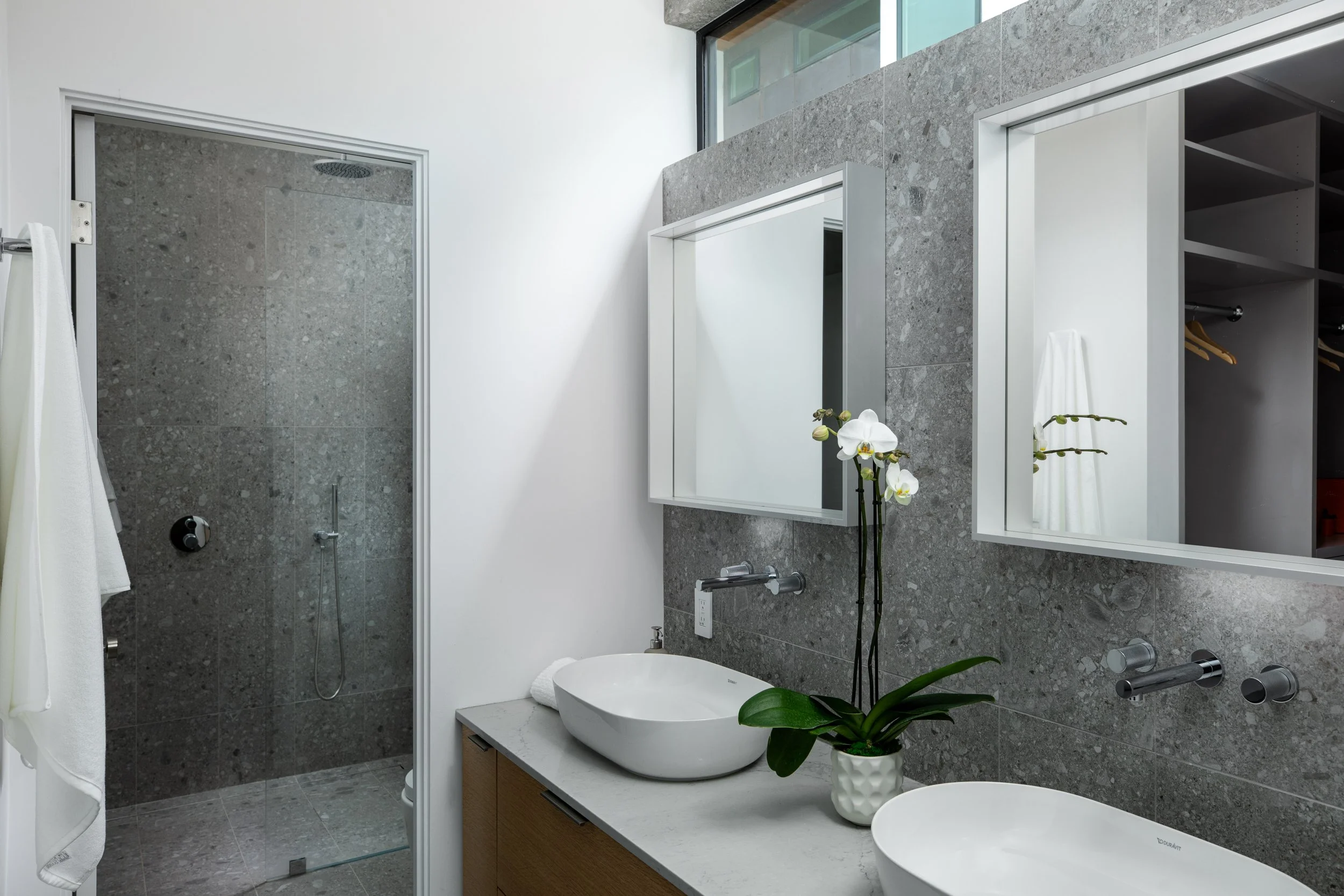 Modern bathroom with gray tiled walls, a walk-in shower with a glass door, a white vessel sink, a mirror, and a potted orchid on the countertop.