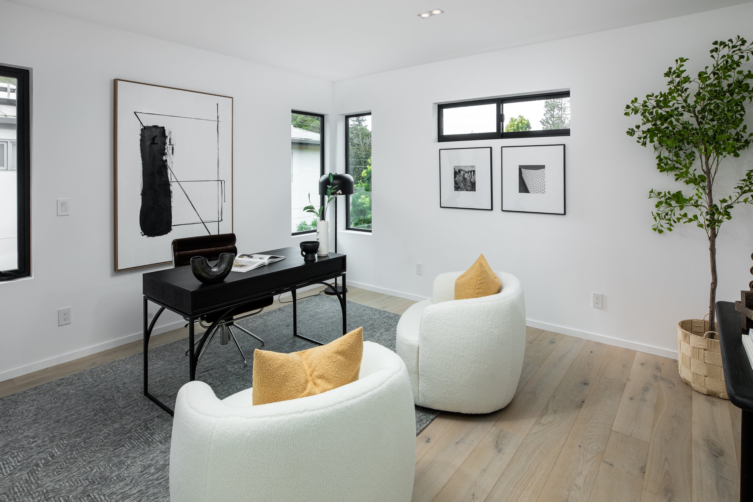 Modern minimalist living room with white walls, light wood flooring, and large windows. Features a black desk with decorative items, two white armchairs with yellow pillows, framed black-and-white art on the walls, a tall indoor tree in a woven baske