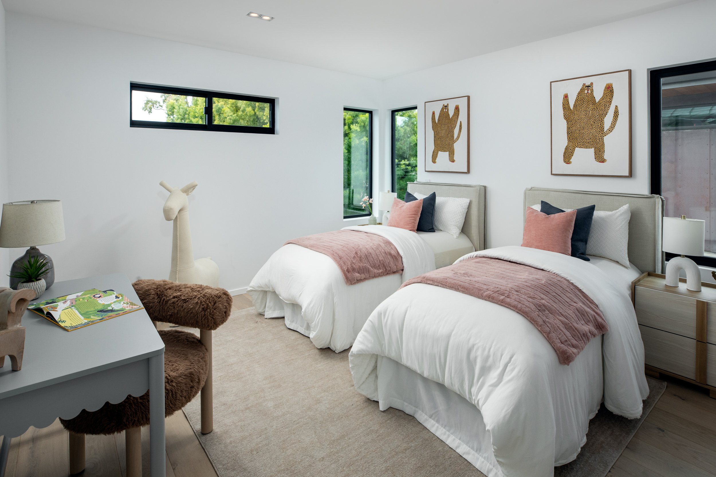 A cozy bedroom with two twin beds with beige headboards, white bedding, and pink and navy pillows. There are three framed pictures of leopardabove the beds. The room has a beige carpet, light wood side tables, and a window with a view of green trees.