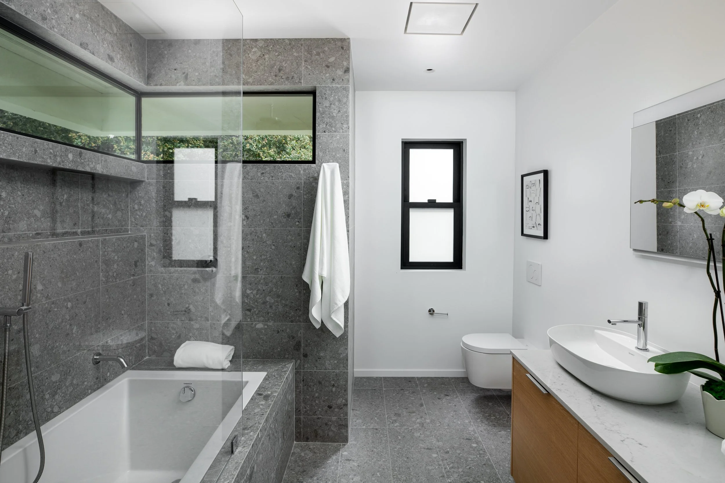 Modern bathroom with gray tiled walk-in shower, bathtub, white walls, black-framed window, wall-mounted toilet, white vessel sink on wooden vanity, and a framed artwork on the wall.