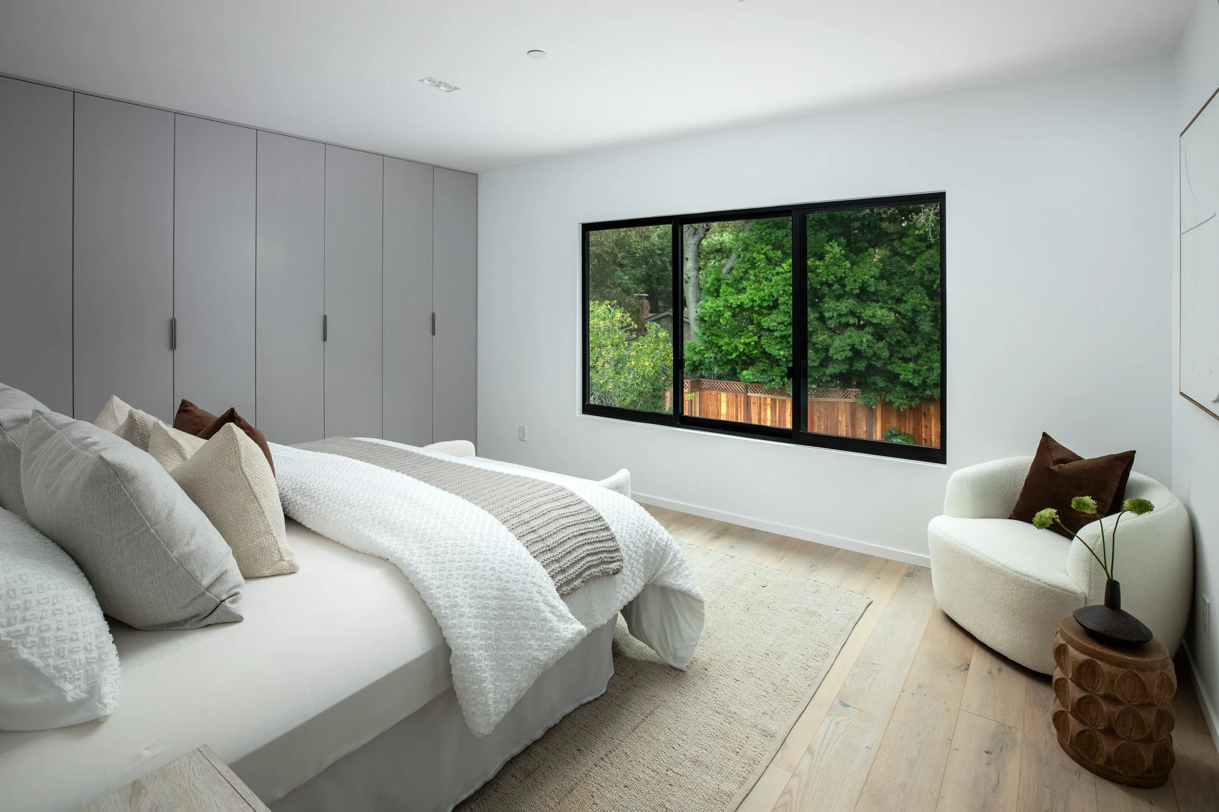A modern bedroom with a white bed, gray pillows, beige and brown pillows, a large window overlooking trees and a wooden fence, a cozy cream chair with a brown pillow, a small wooden side table with a black vase and green flowers, a light-colored rug,