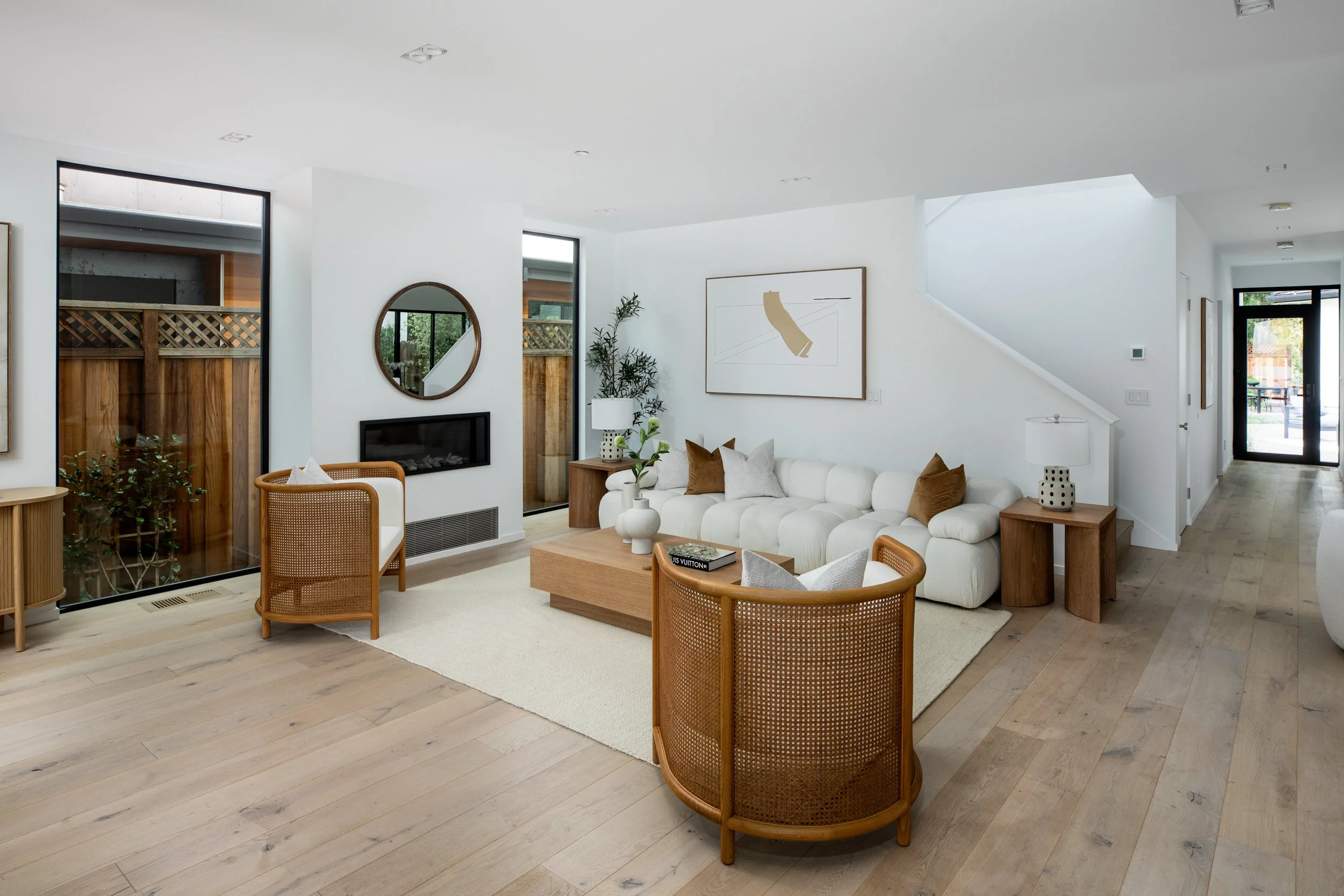 Modern living room with a white sofa, wooden chairs, greenery, framed artwork, and a fireplace.