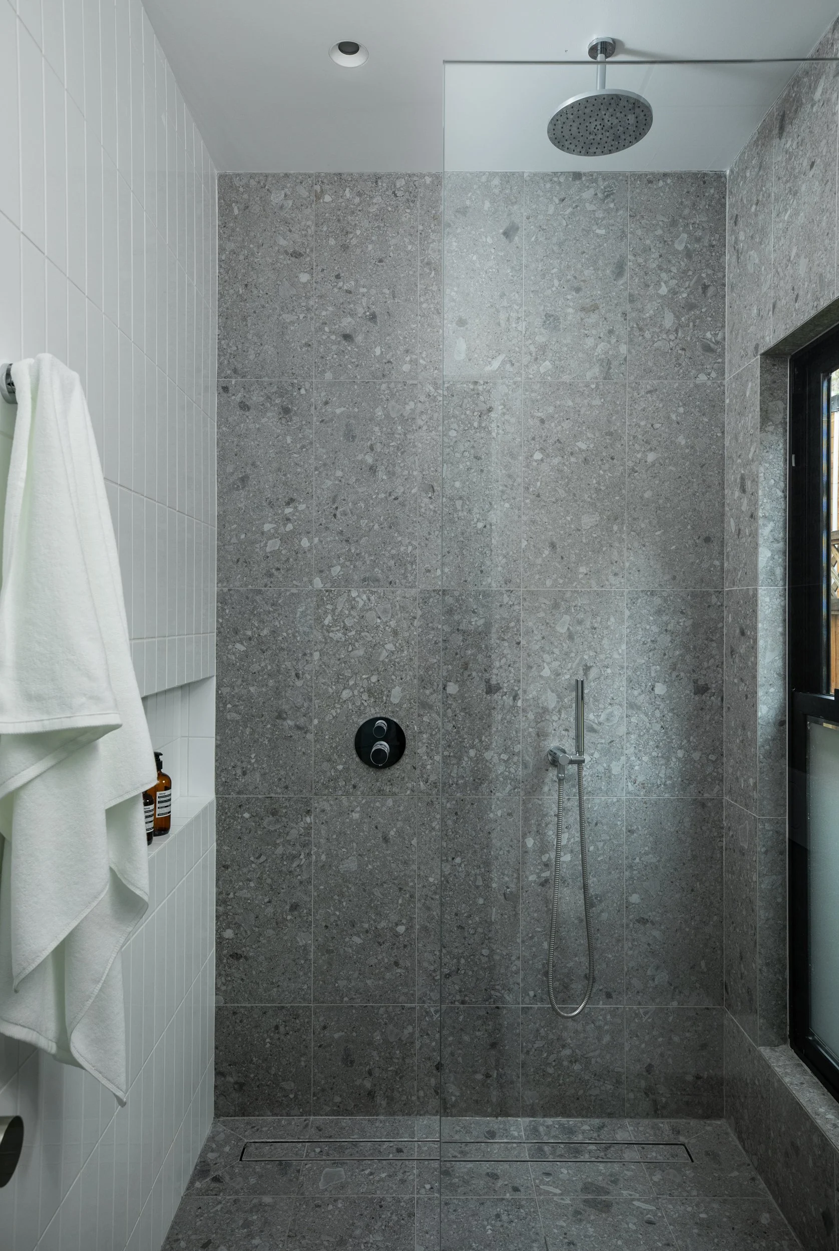 Modern shower area with grey textured wall tiles, a round rain shower head, a black control panel, a handheld shower head, white towel hanging on a hook, and a small window with black frame on the right side.
