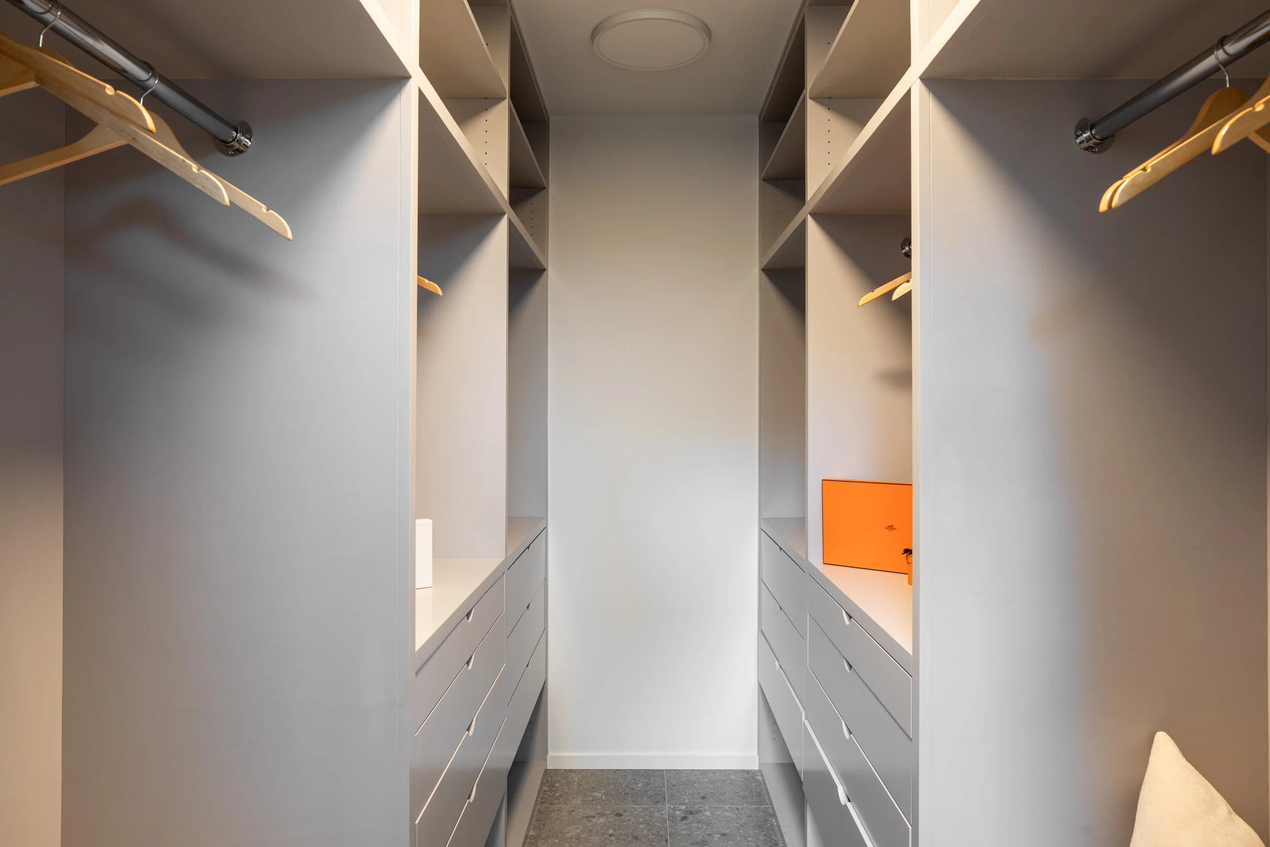 Empty walk-in closet with white built-in shelves and drawers on both sides, and clothing rods with wooden hangers. The floor is dark grey tile, and a small orange box is on the right side shelf.