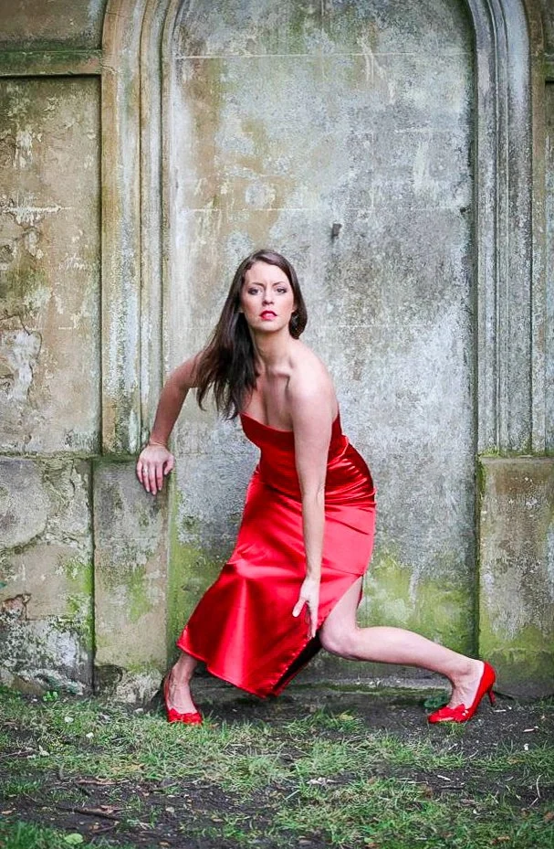 A woman in a red satin dress and matching red high heels is posing outdoors in front of a weathered stone wall, with her left leg bent and her right leg extended in a lunge position.