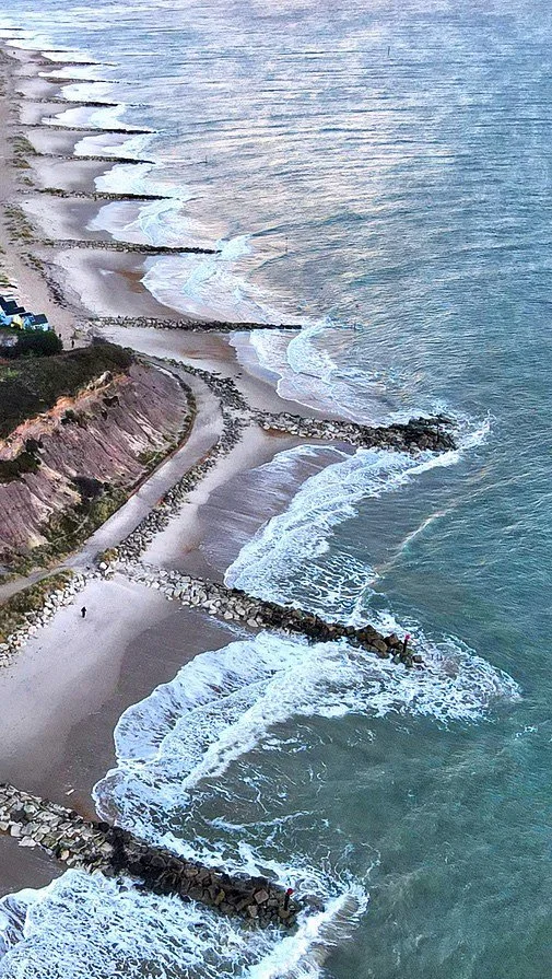 Aerial view of a beach with multiple rock breakwaters extending into the ocean, waves crashing against them, and a sandy shoreline with a few people and a rocky cliff on the side.