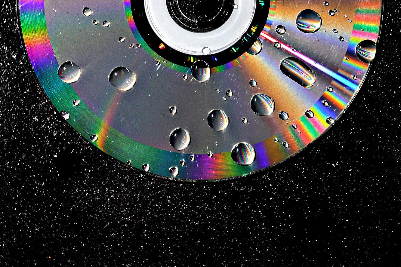 Close-up of a CD disc reflecting rainbow colors with water droplets on its surface, set against a black starry background.