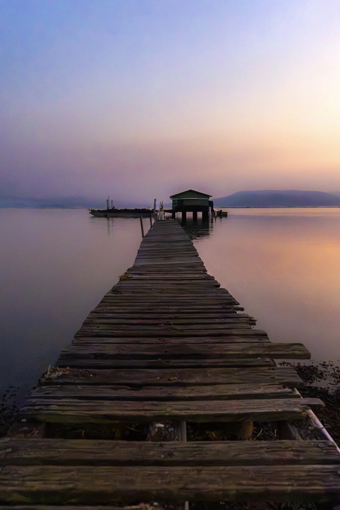 A wooden dock extends into calm water, leading to a small structure over the water at sunset with a pastel sky and distant mountains.