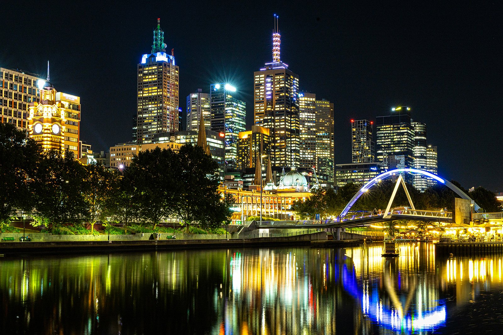 Night view of a city skyline with illuminated skyscrapers, a river with reflections, and a bridge with blue lights.