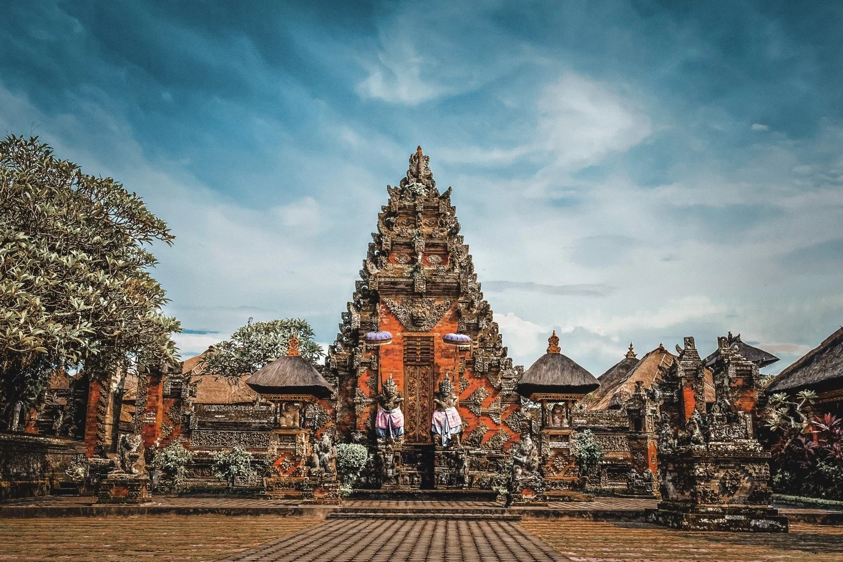An ornate traditional Balinese temple with intricate carvings, statues, and thatched roofs, set against a partly cloudy sky with surrounding trees.