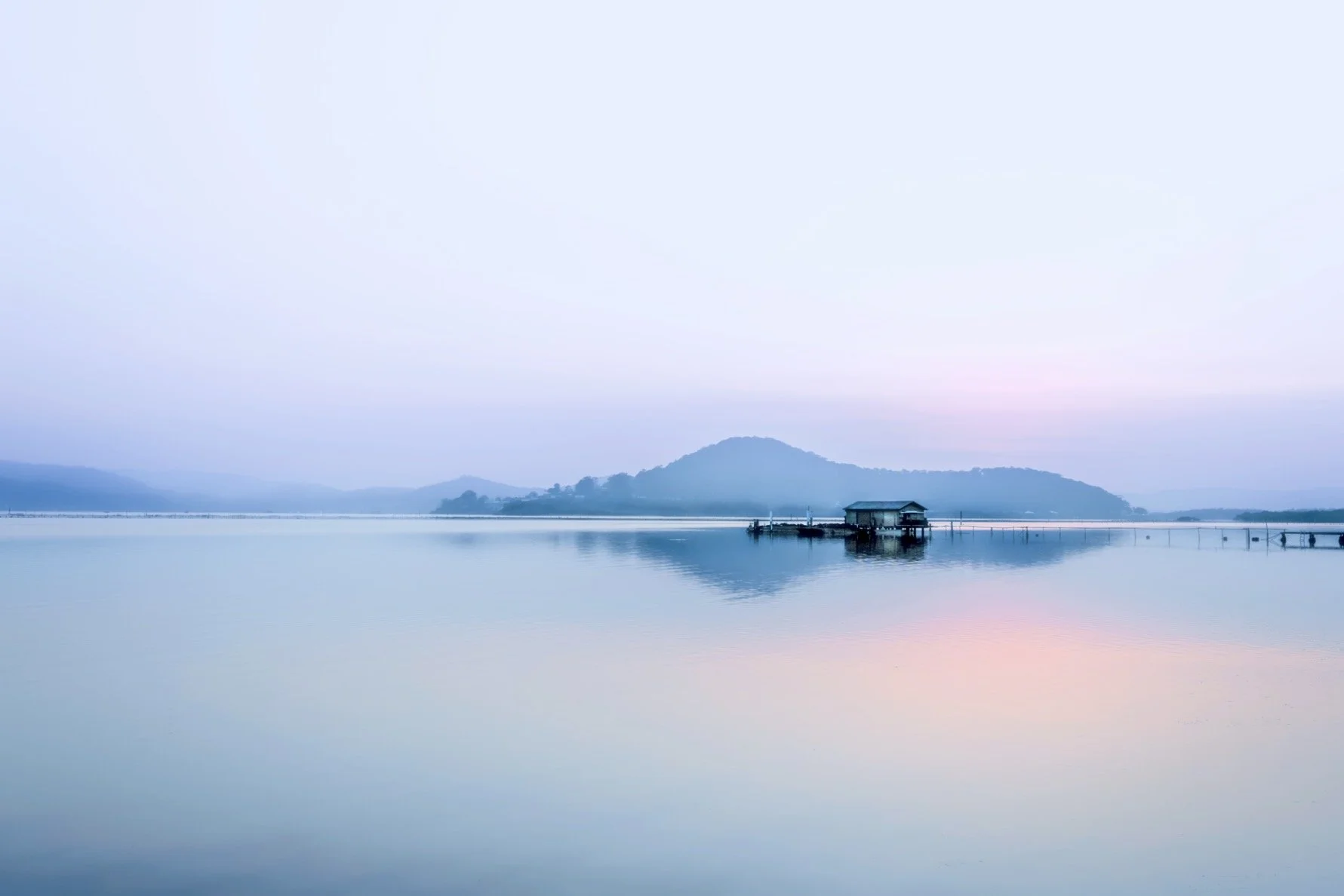 Calm lake with mountains in the background, a small pier and house on stilts, reflecting serene pastel sky.