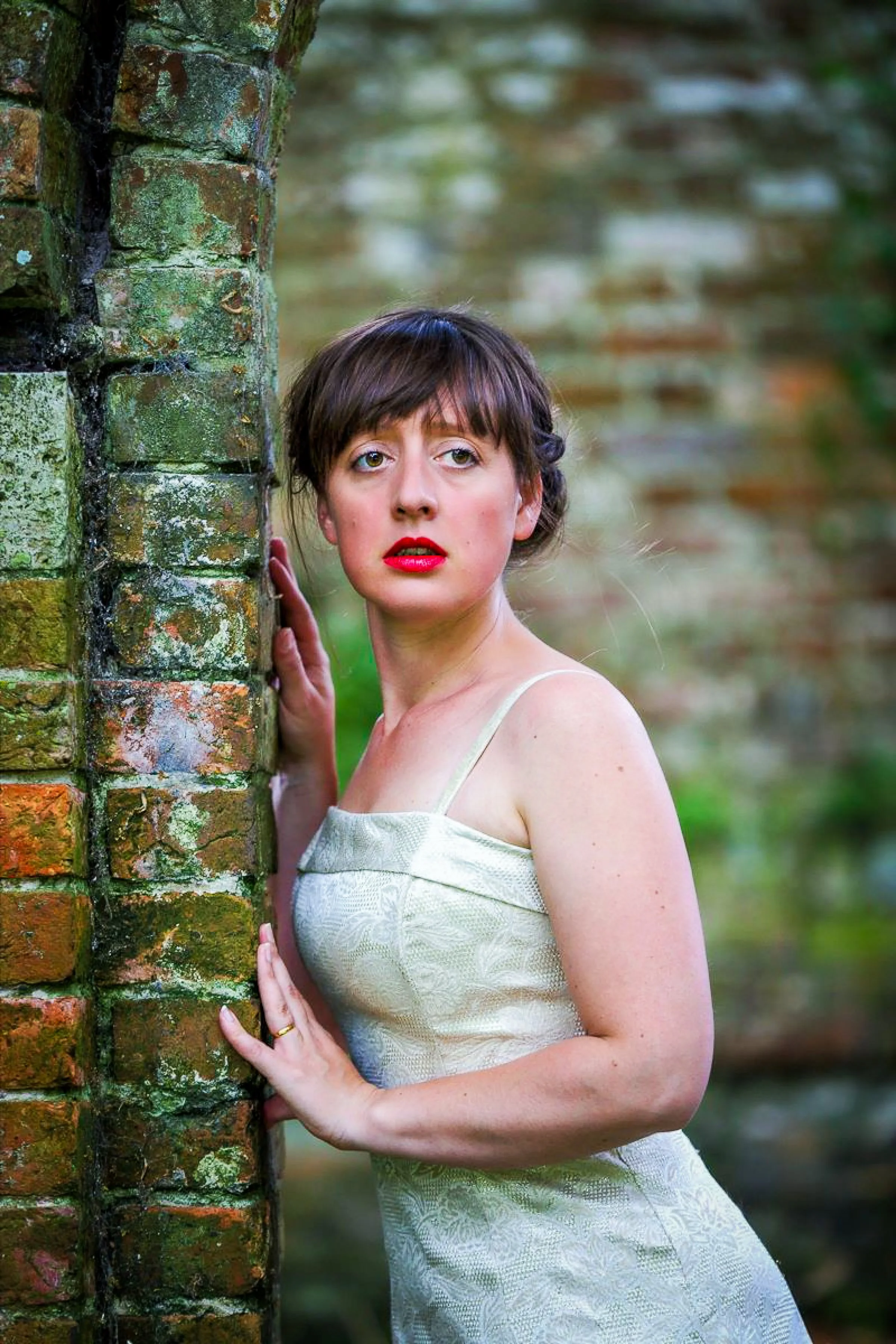 A woman with short brown hair, wearing a light-colored dress, holding onto a moss-covered brick wall and looking into the distance.