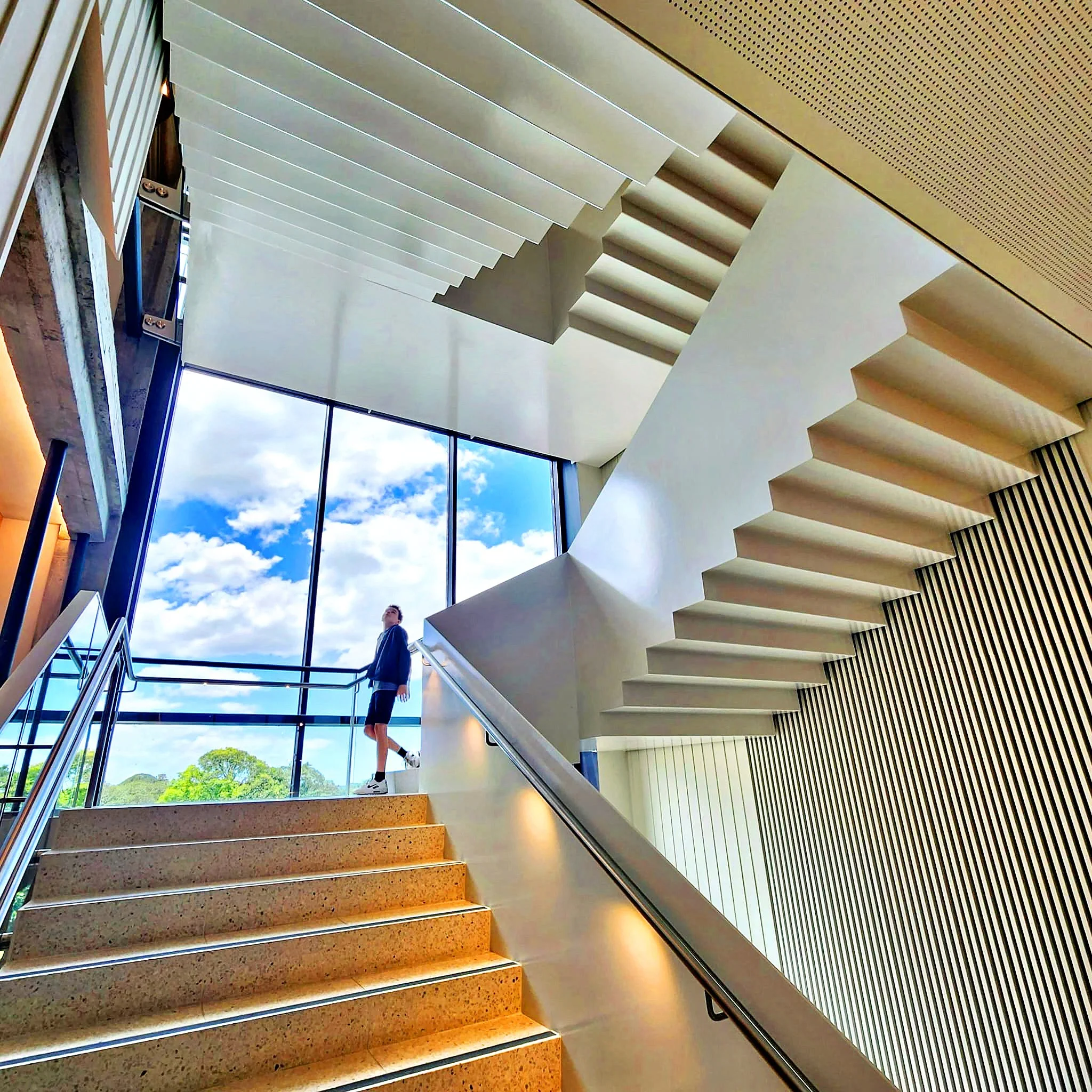 Modern indoor staircase with a person standing at the top, large floor-to-ceiling windows showing a blue sky with clouds and green trees outside.