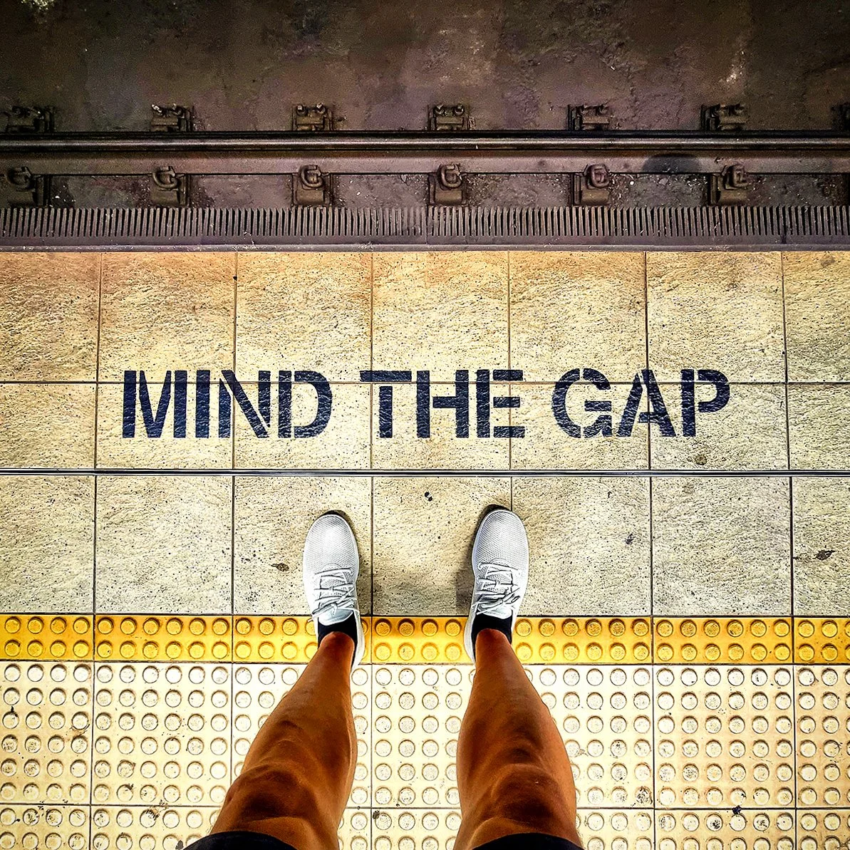View of a subway platform with a person standing, wearing white shoes and shorts, looking down at the tile floor which has the words "MIND THE GAP" painted on it in large black letters.