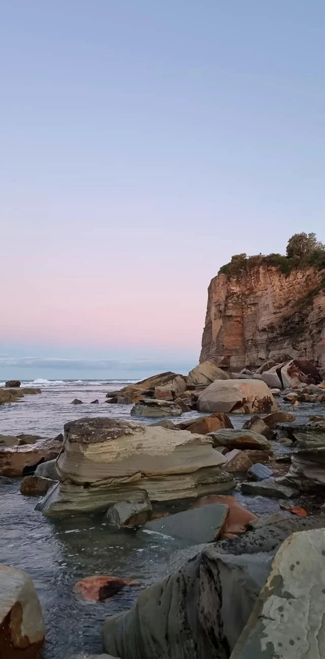 Rocky coastline with large rocks in shallow water, tall cliff on the right, and a clear sky with soft pastel colors in the background.