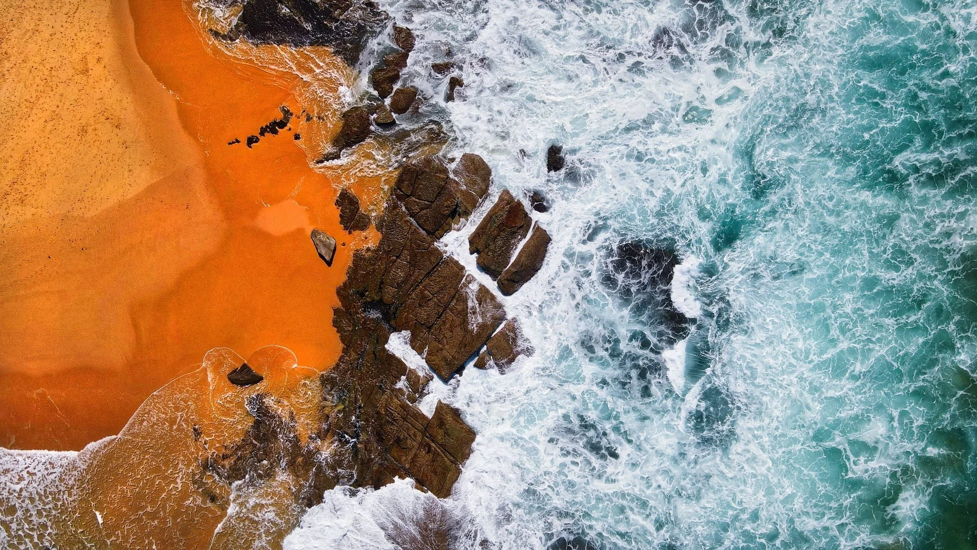Aerial view of a rocky shoreline with waves crashing onto orange sandy beach.