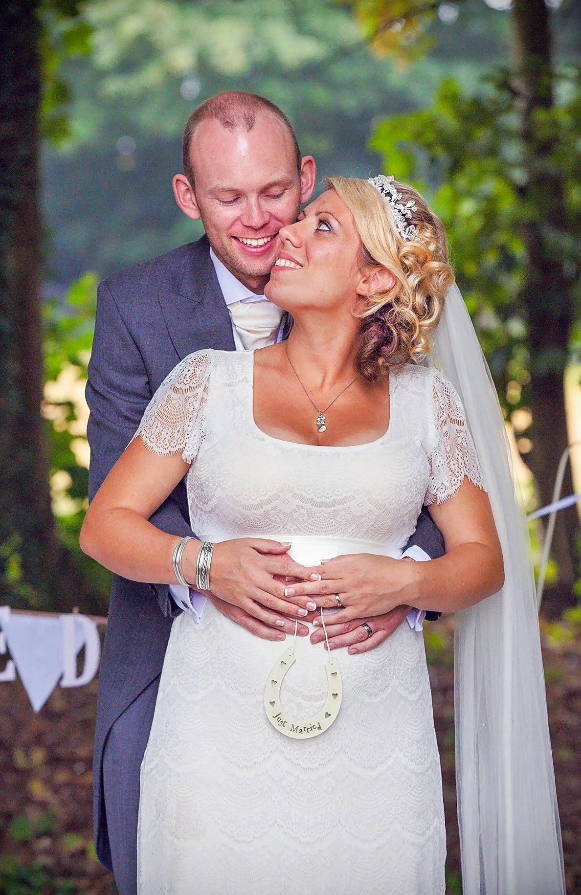 A happy bride and groom are embracing outdoors, with the groom standing behind and holding the bride's waist. The bride wears a white lace wedding dress and a veil, with a horseshoe-shaped ornament hanging from her waist that reads 'Just Married.' Th