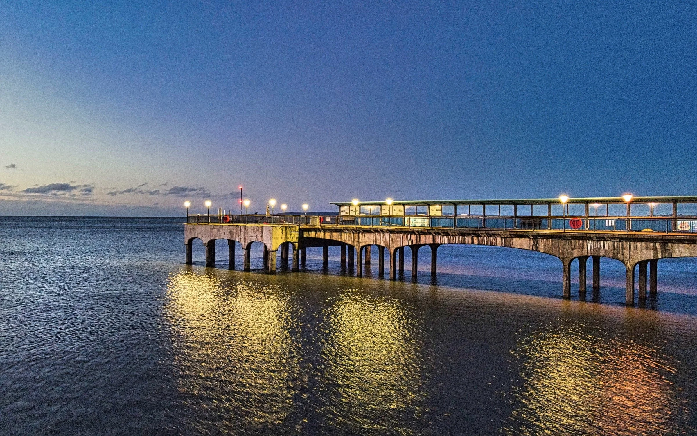 A pier extending into the water at sunset, with the sky transitioning from light to dark and the pier lights reflecting off the calm water.
