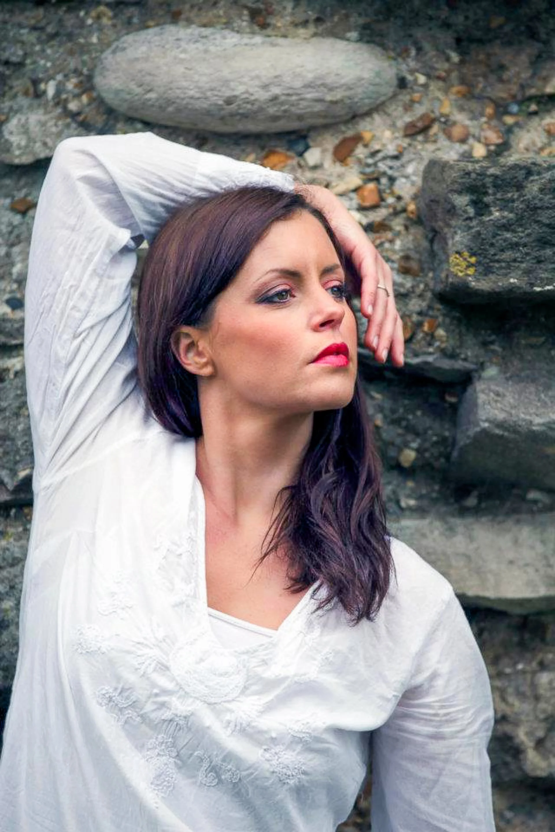 A woman with dark hair and makeup, wearing a white embroidered top, is posing with one arm raised behind her head against a rocky background.