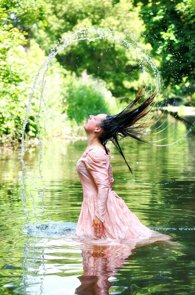 A woman in a pink dress standing in a body of water, flipping her wet hair back, creating an arc with water droplets in a lush green outdoor setting.