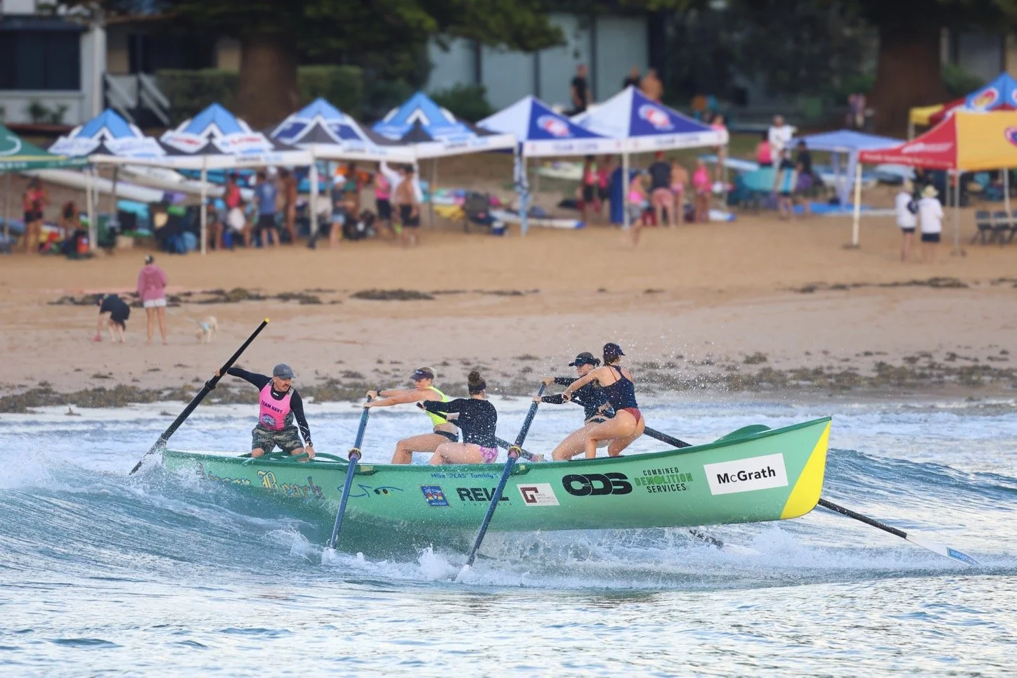 Four women in swimsuits paddling a green canoe on the water near a beach with people and tents in the background.