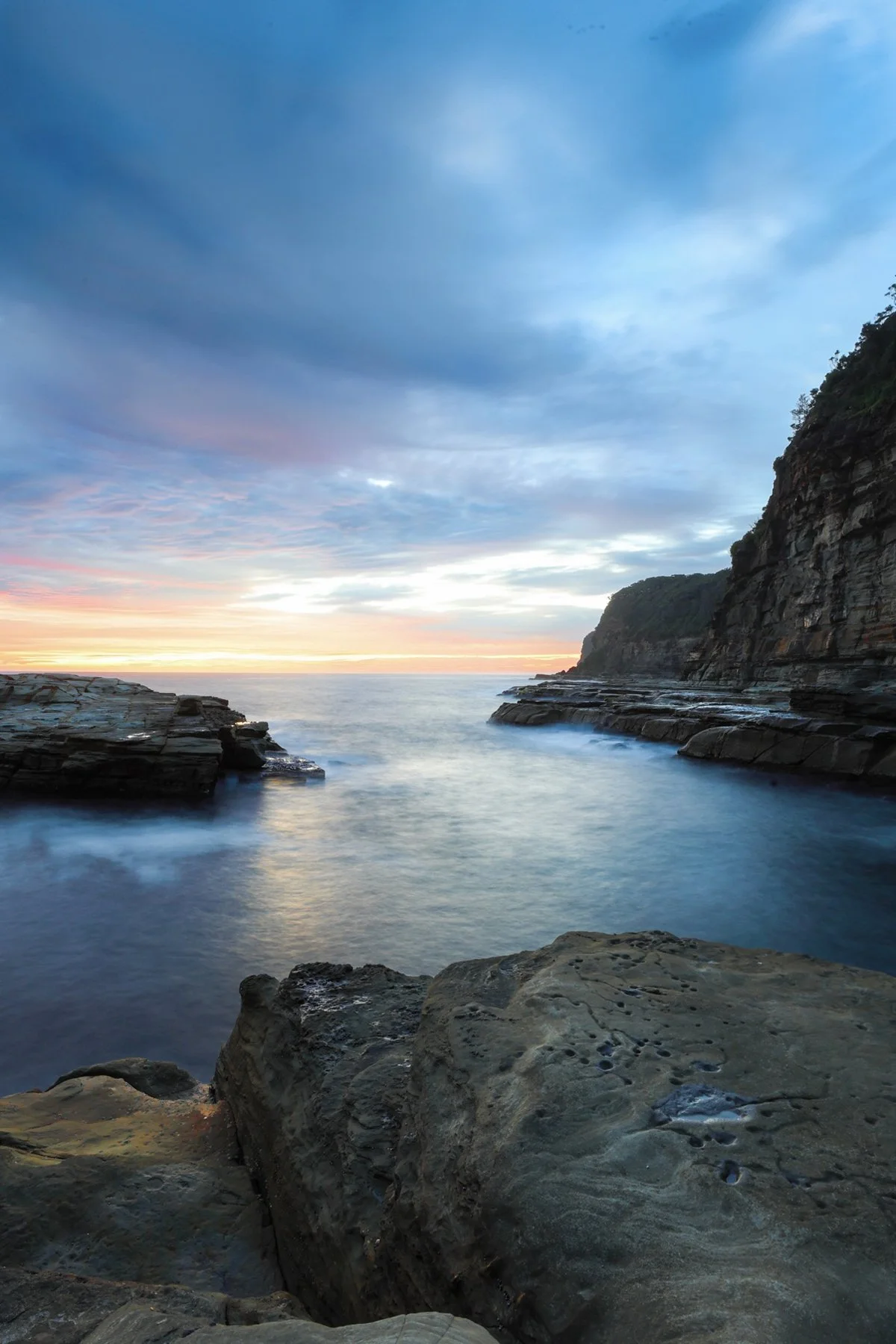 Scenic coastal view at sunset with rocky shoreline, calm water, cliffs, and a colorful sky.