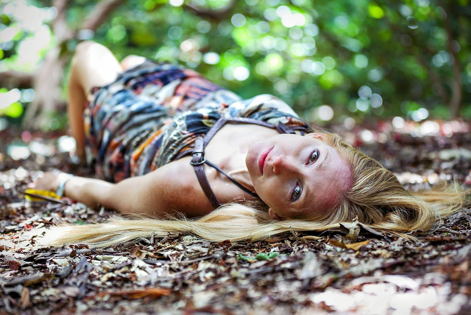 A woman with long blonde hair lying on the forest floor covered in fallen leaves, with trees and greenery in the background.