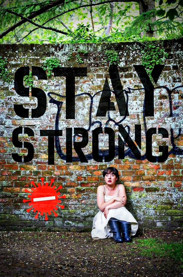 A young woman sitting on the ground against a brick wall with the words 'STAY STRONG' painted over it. The wall has green moss and graffiti. There is a red symbol resembling a virus with a white bar across it near the bottom left.