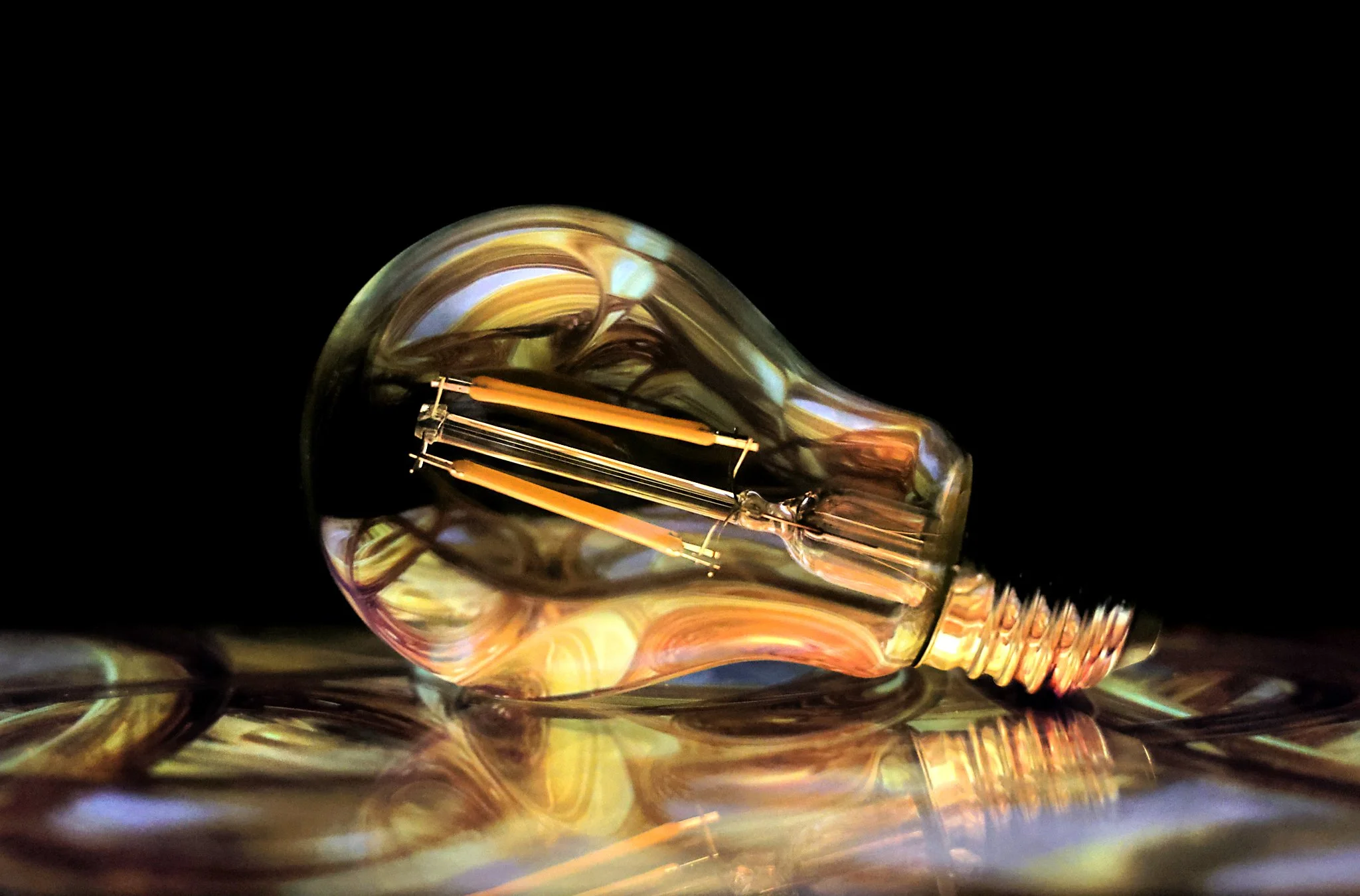 Close-up of an illuminated vintage incandescent light bulb lying on a reflective surface against a black background.