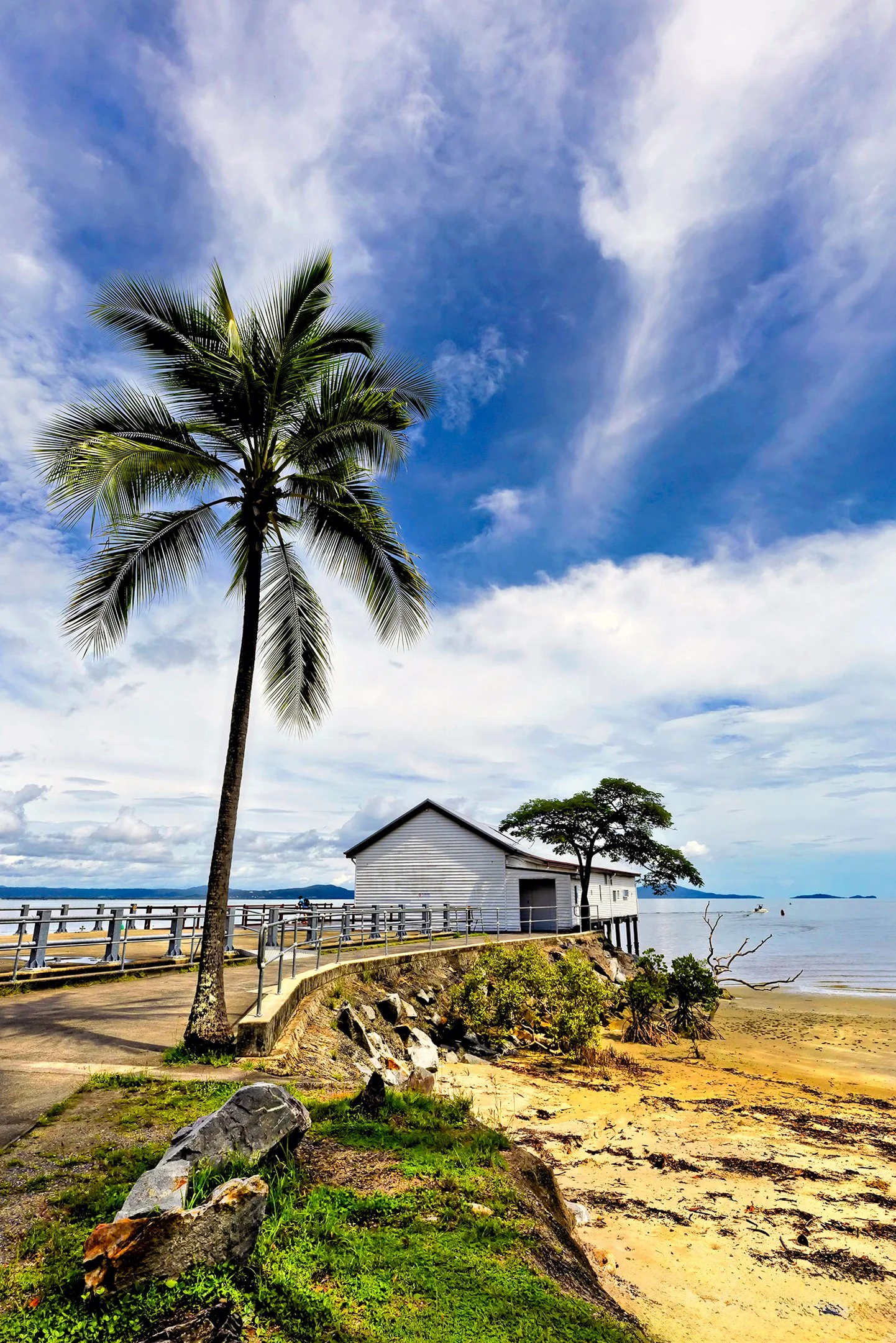 A tropical beach scene with a large palm tree, a white wooden building on stilts, sandy shore, ocean water, and a partly cloudy blue sky.