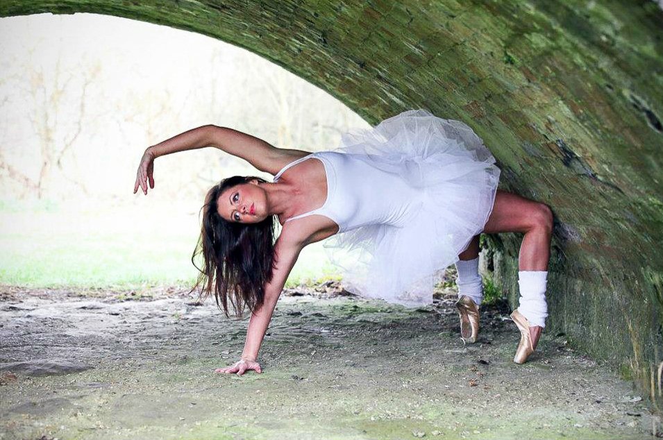 A female ballet dancer in a white tutu and ballet slippers performs a dance move while balancing on her hands and feet under a stone bridge.