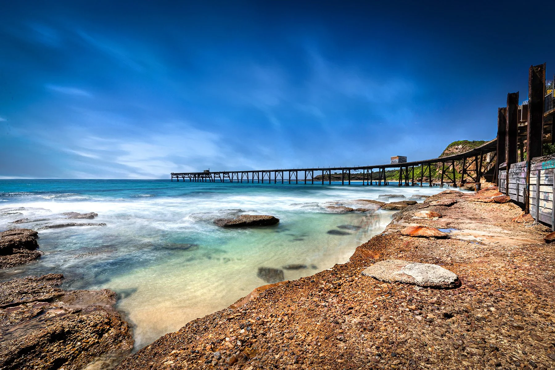 View of a rocky beach with a long pier extending into the ocean under a blue sky.