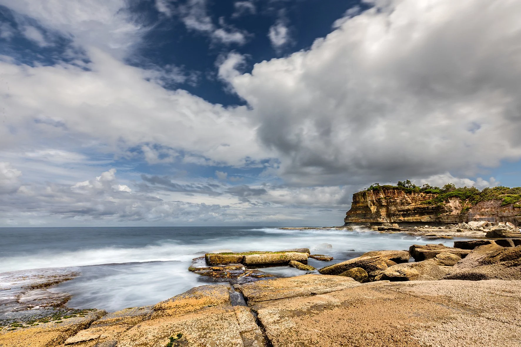 Scenic coastal view with sandy beach and rocky shore, presenting a cliff in the distance under a partly cloudy sky.