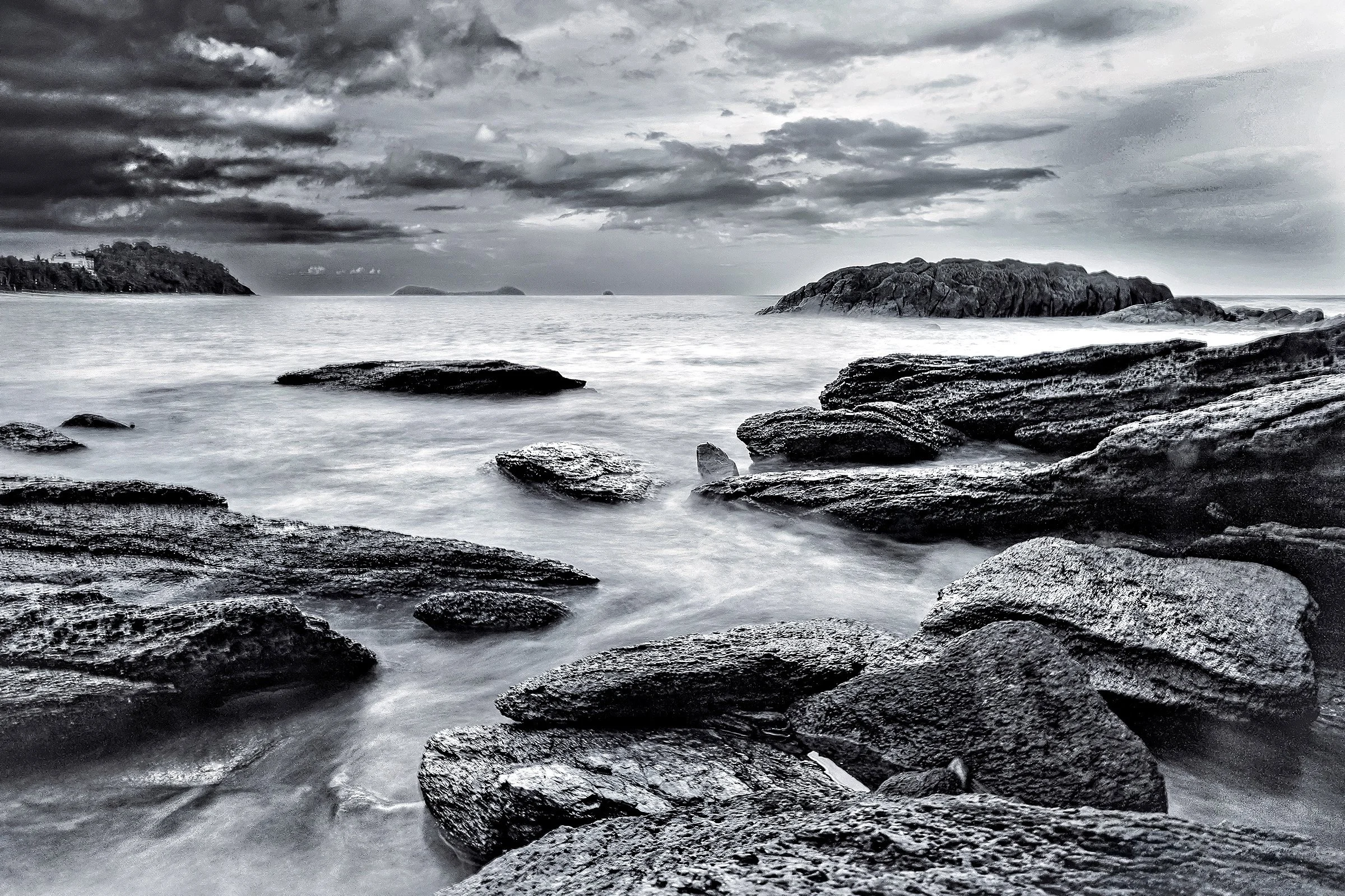 Black and white photograph of rocky shoreline with sea waves, cloudy sky, and distant islands.