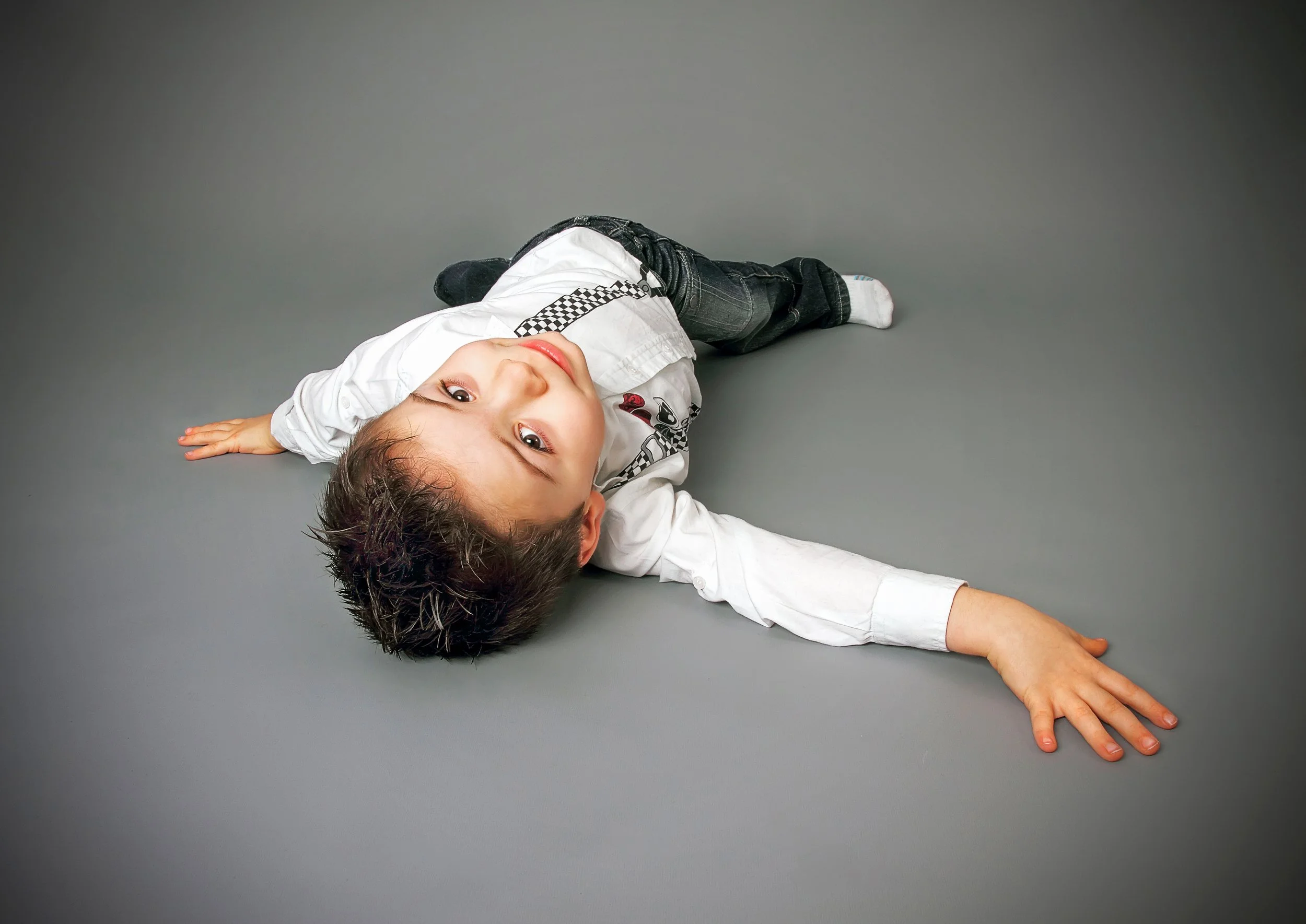 A young boy with dark hair lying on the floor with one arm extended and the other bent, looking up at the camera with a playful expression, on a gray background.