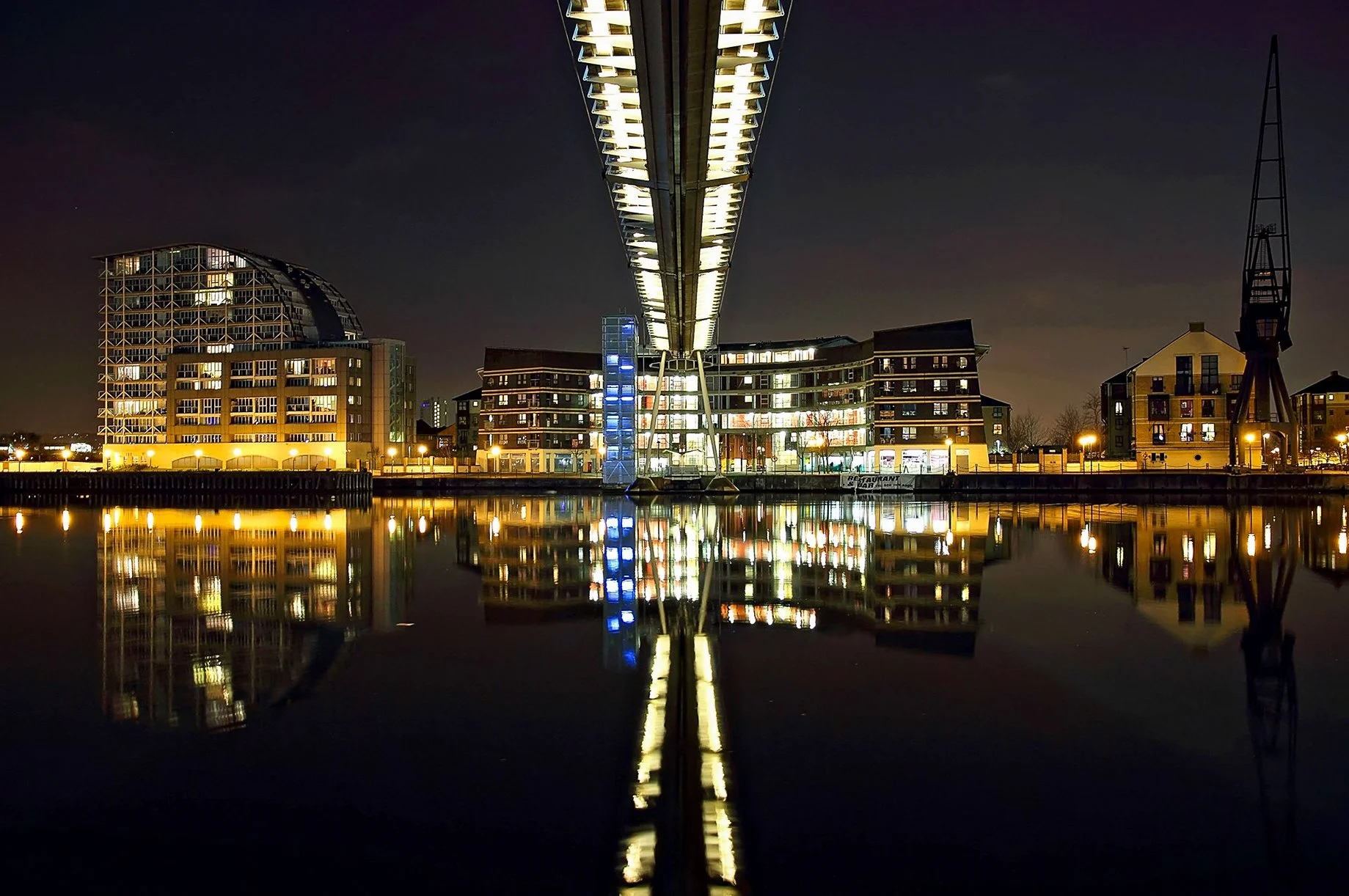 Night view of modern buildings reflected in calm water with a bridge overhead, illuminated with lights, in an urban area.