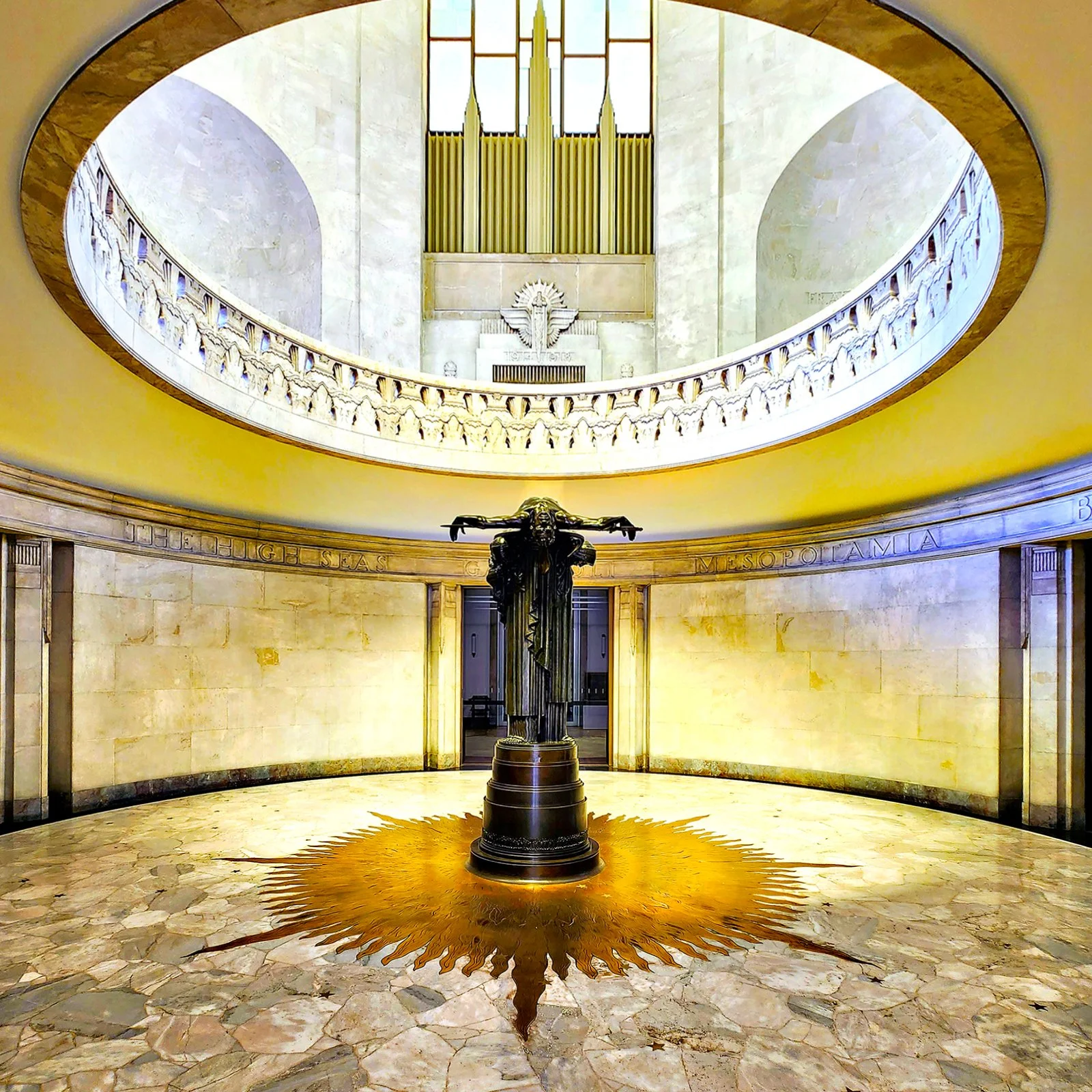 Interior of a church or cathedral with a statue of Jesus Christ on the cross in the center, a sunburst design on the floor beneath it, and an organ with pipes and a dove symbol above in the background.