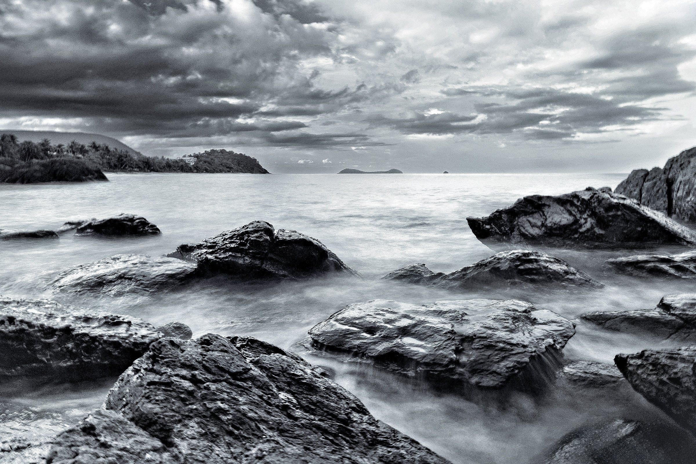 Black and white photo of a rocky shoreline with water and distant islands, under a cloudy sky.