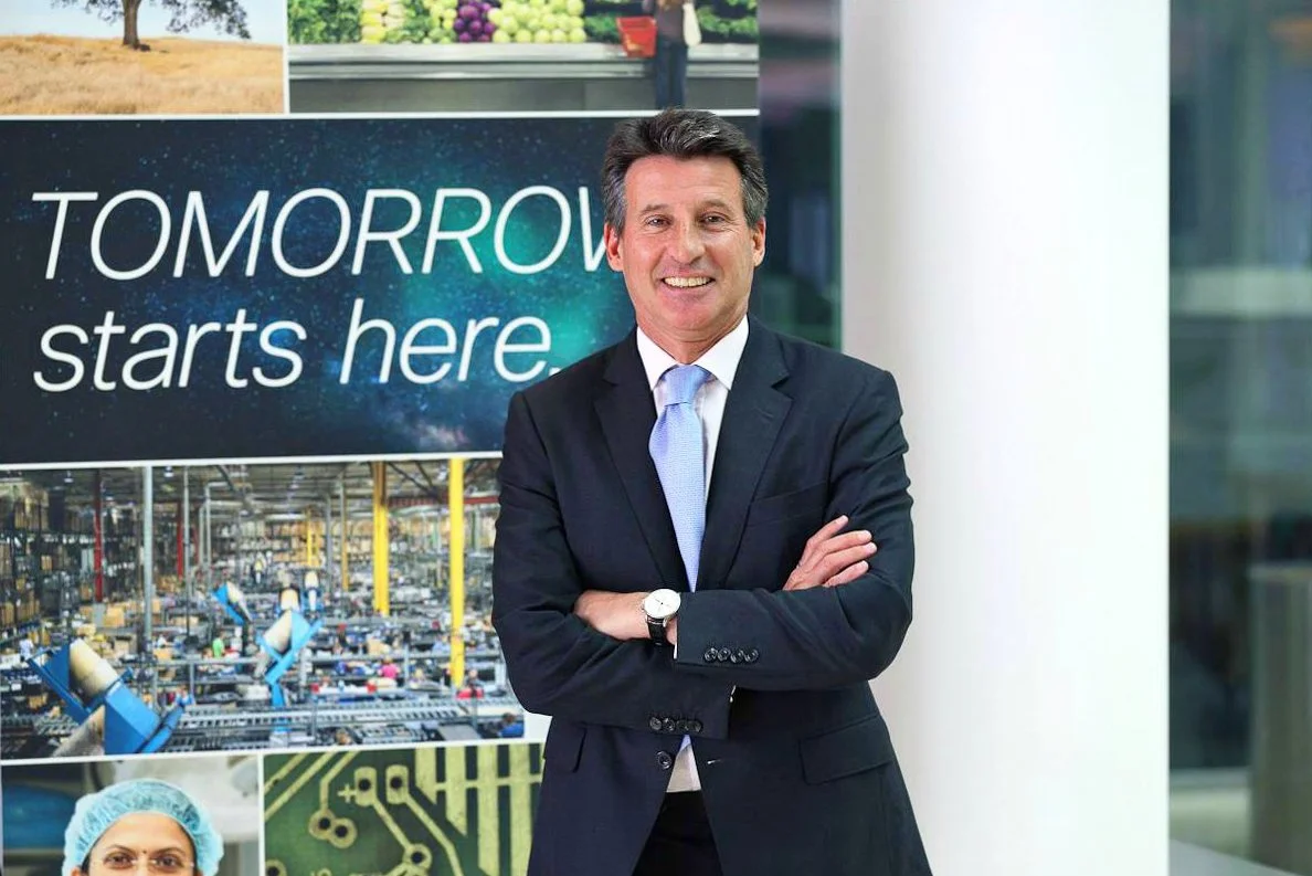 A man in a dark suit and light blue tie standing with arms crossed in front of a banner that reads "TOMORROW starts here". The background includes images of a farm, a flower market, a manufacturing plant, a scientist, and a circuit board.