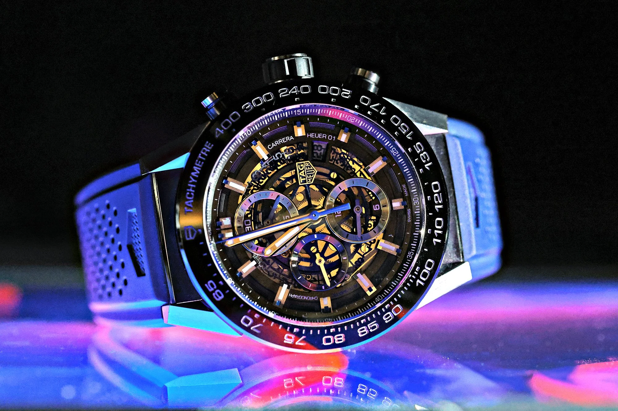 A close-up of a TAG Heuer wristwatch with a skeleton dial, black case, tachymeter bezel, and a blue strap, illuminated with colorful lighting.