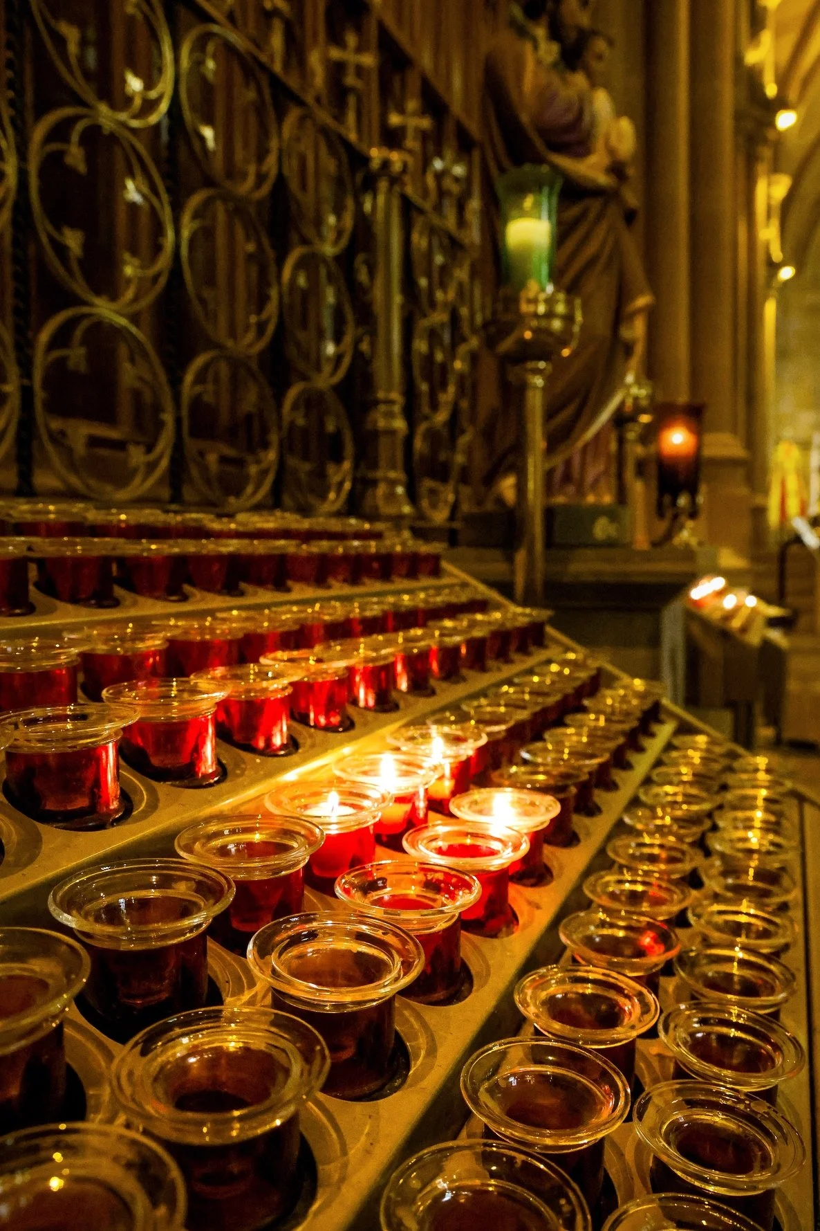 Numerous small glass cups filled with red liquid on a gold-colored stand inside a dimly lit church or cathedral, with gothic-style metalwork, candles, and religious statues in the background.