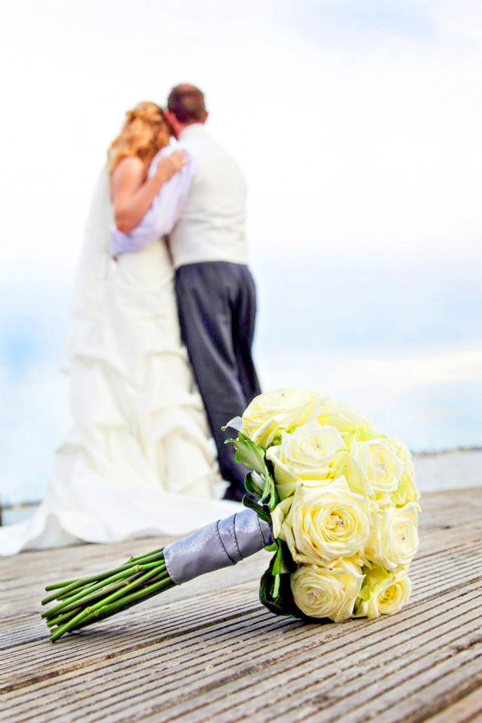 Wedding bouquet of white roses on wooden surface with a bride and groom kissing in the background.