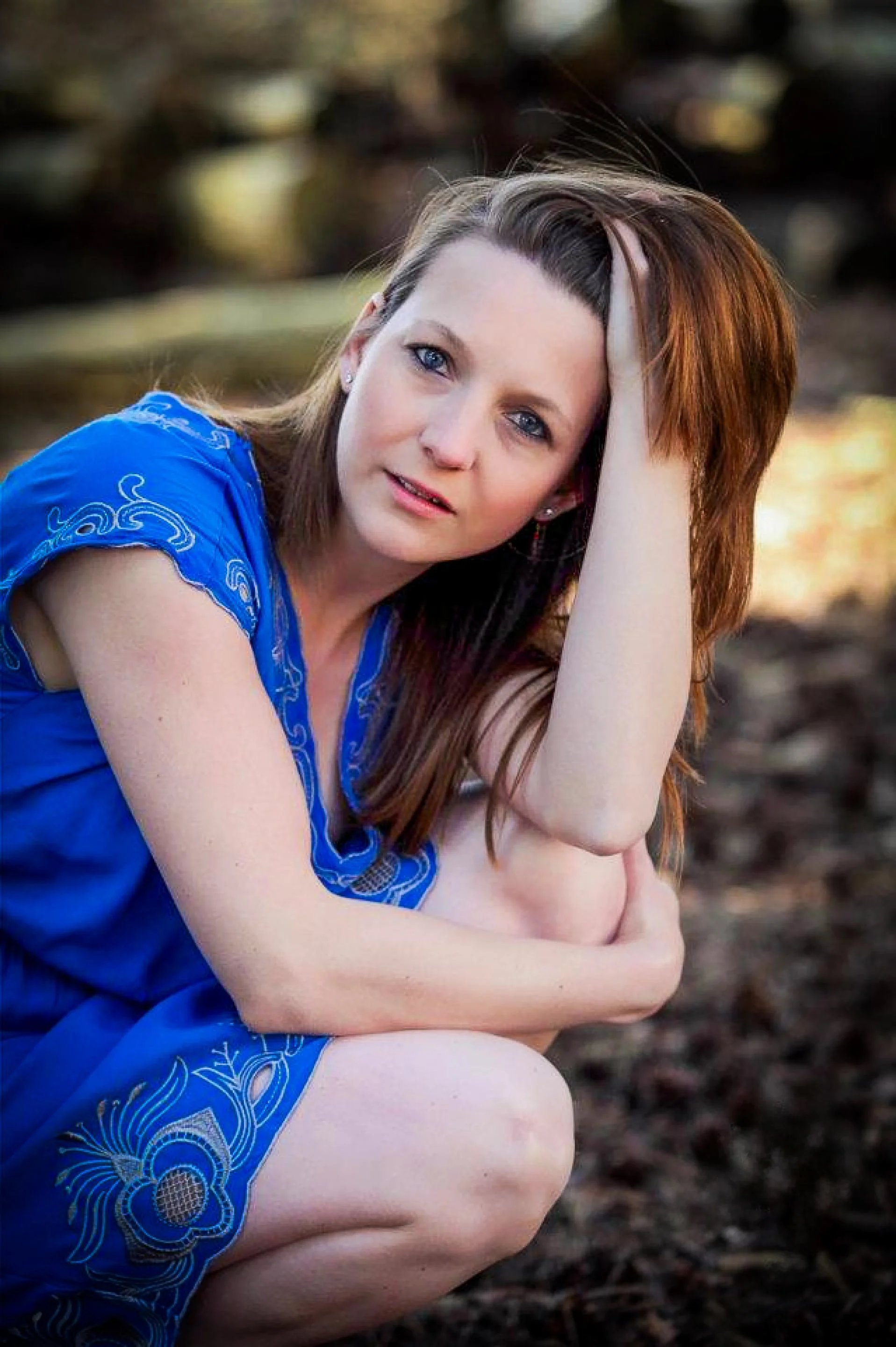 A woman with red hair, wearing a blue dress, crouching outdoors with one hand on her head, looking at the camera with a pensive expression.