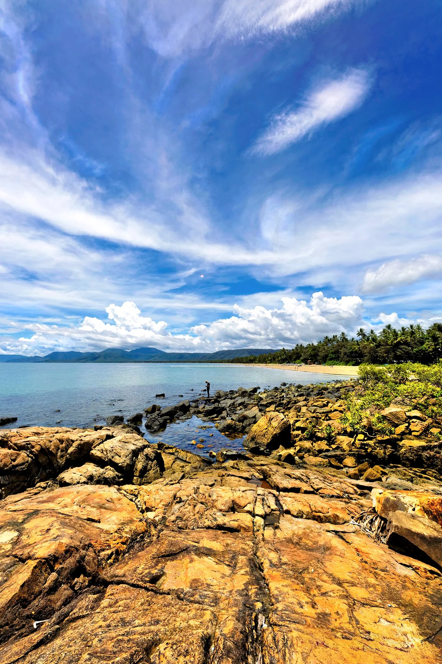 A scenic coastal landscape with rocks in the foreground, calm water, and a person fishing. The background features lush greenery, a sandy beach, migration clouds in a bright blue sky with scattered clouds, and distant mountains.