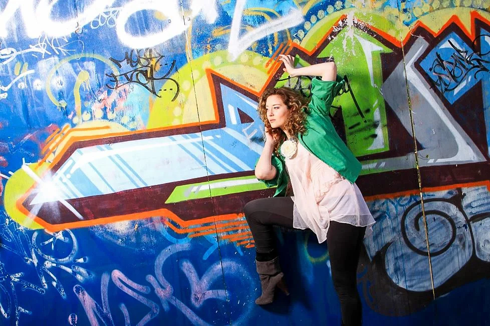 A woman with curly hair wearing a green jacket, white blouse, and black pants is posing in front of a colorful graffiti wall.