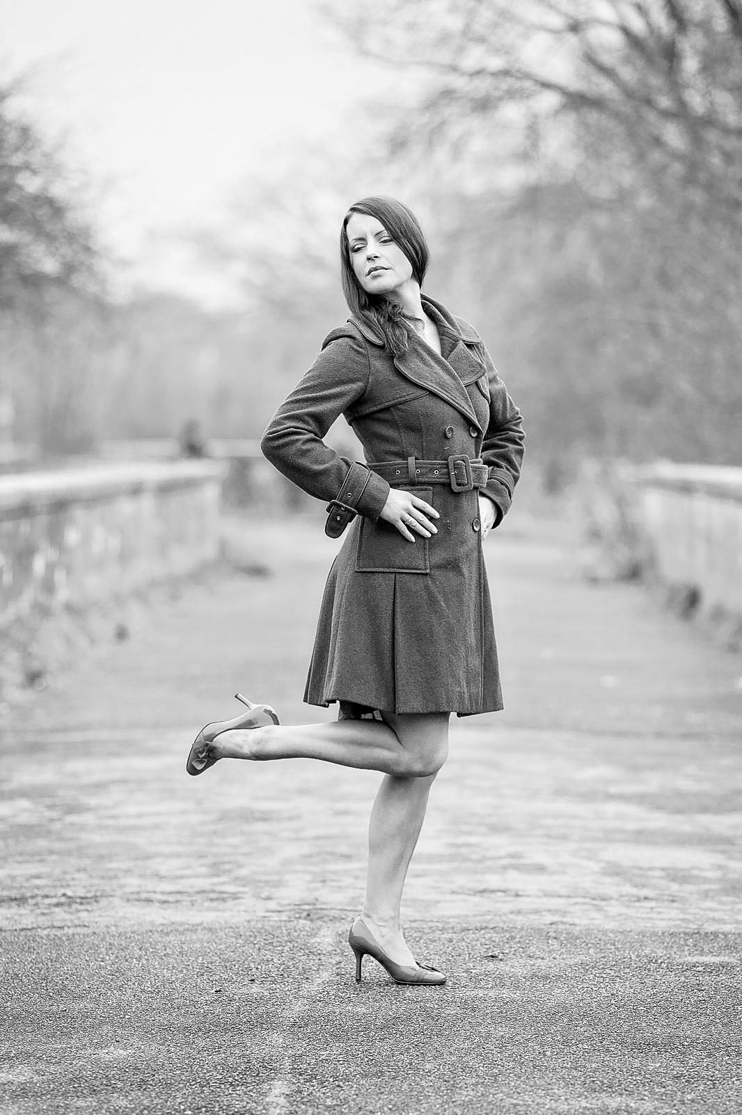 A black and white photo of a woman in a trench coat and high heels standing outdoors on a path with trees in the background, striking a pose with one leg bent backward and her hands on her hips.