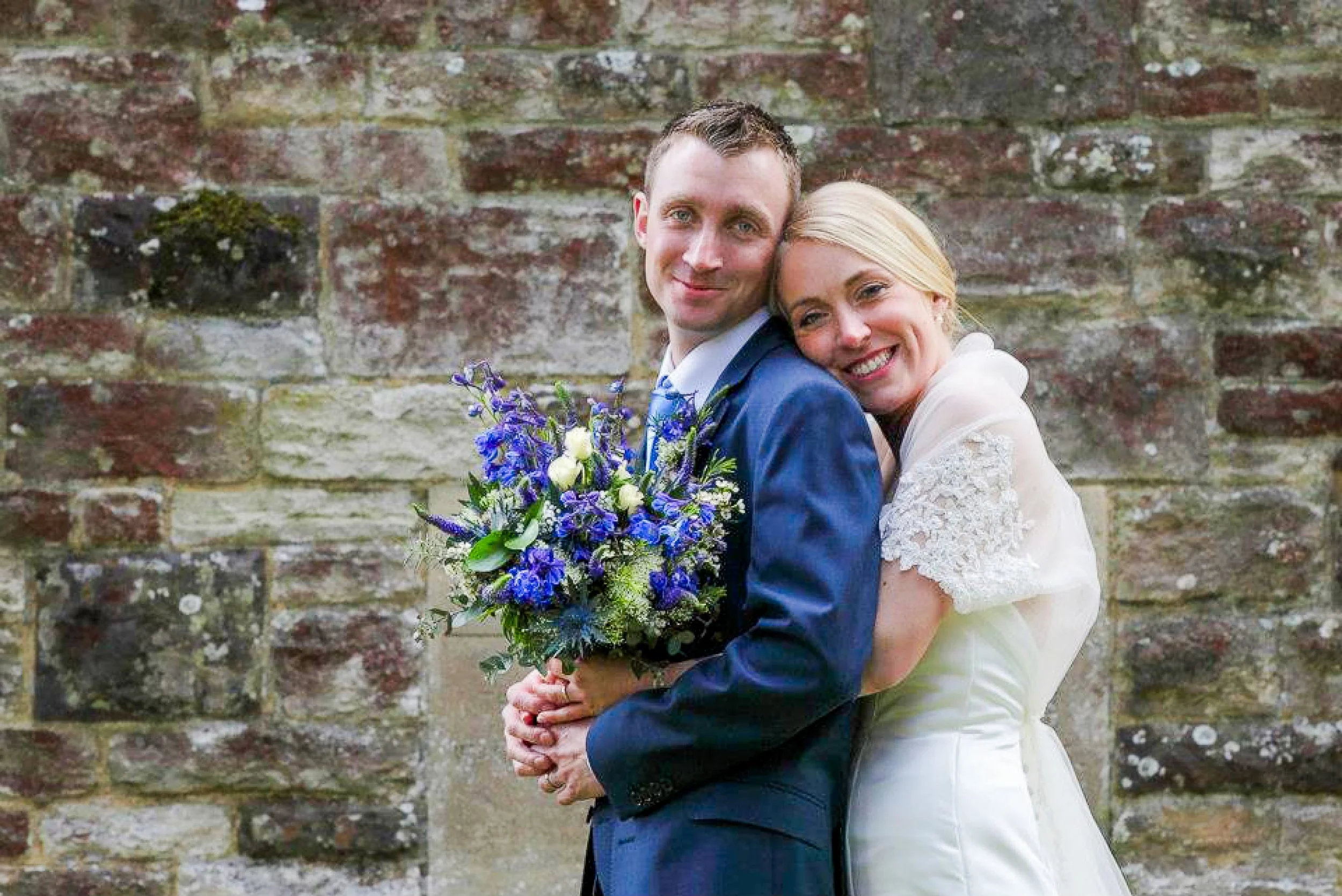 A newlywed couple, the groom in a blue suit and the bride in a white dress, standing in front of a rustic stone wall. The groom is holding a bouquet of blue and white flowers, and they are smiling while embracing.